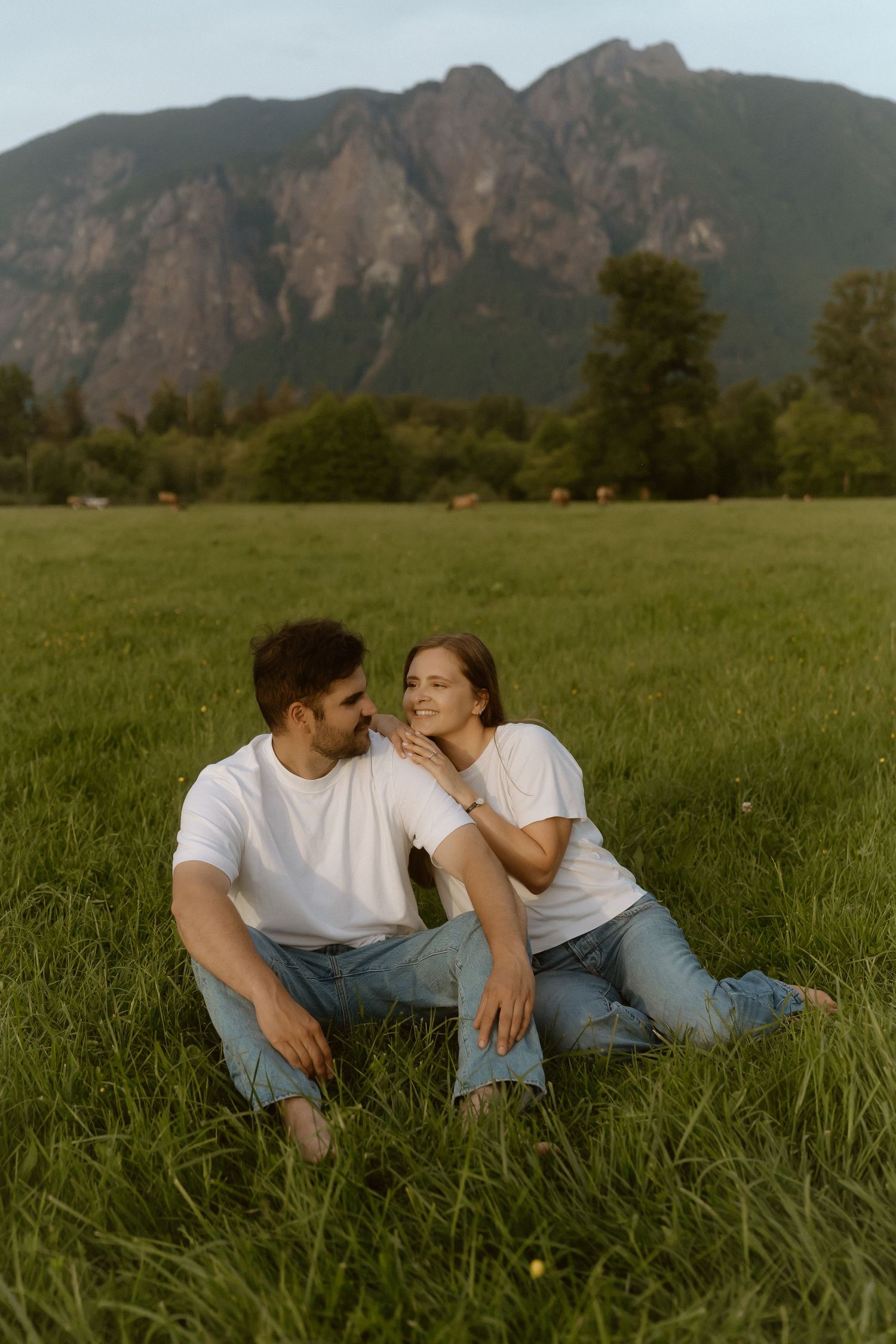 Couple in matching white tees and jeans sitting on green grass field with mountain backdrop.