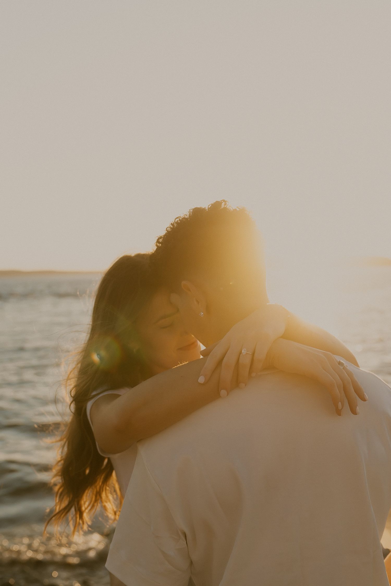 Couple embracing at golden hour on the beach, woman smiling with arms wrapped around her partner as warm sunlight flares over the water.