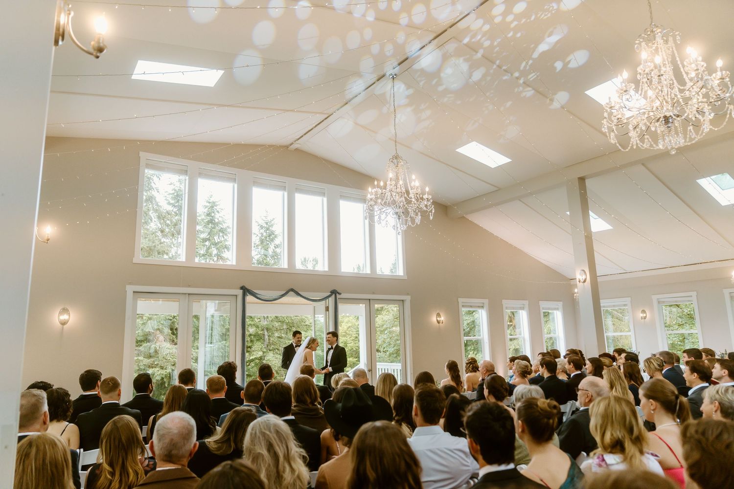 Elegant wedding ceremony in a bright white venue with chandelier, guests seated as couple exchanges vows at the altar.