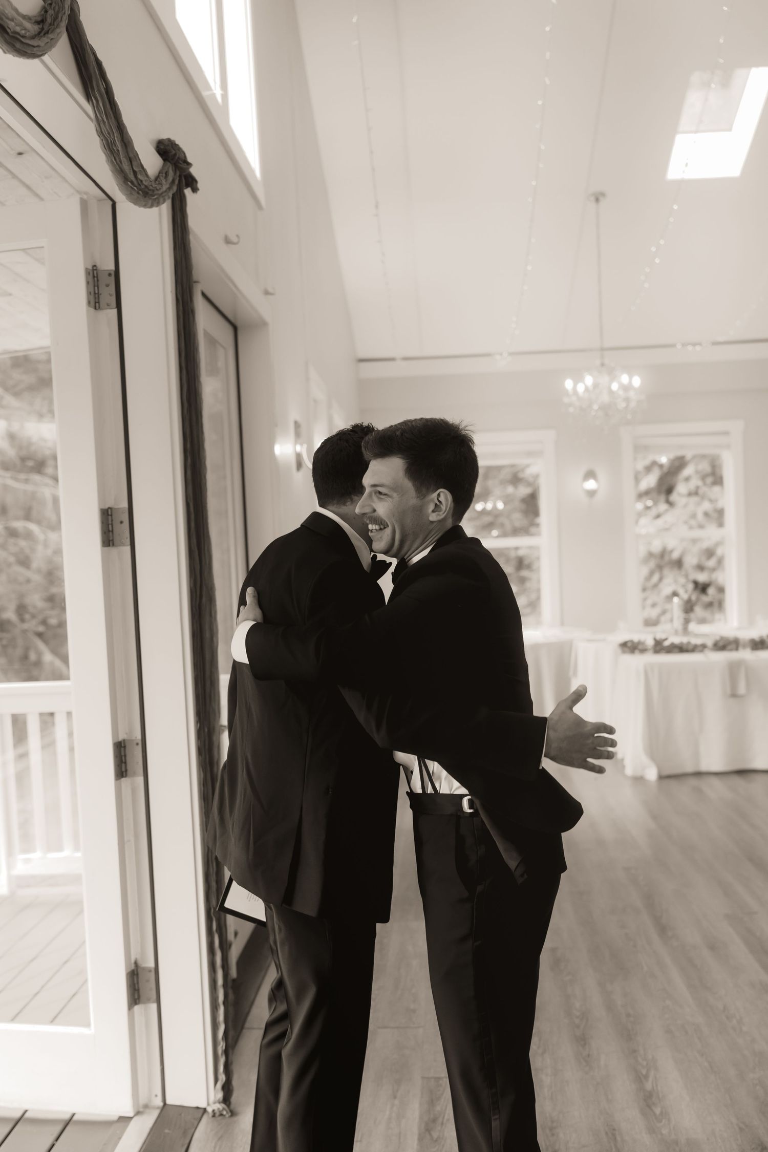 Two grooms in tuxedos share a joyful embrace inside an elegant wedding venue.