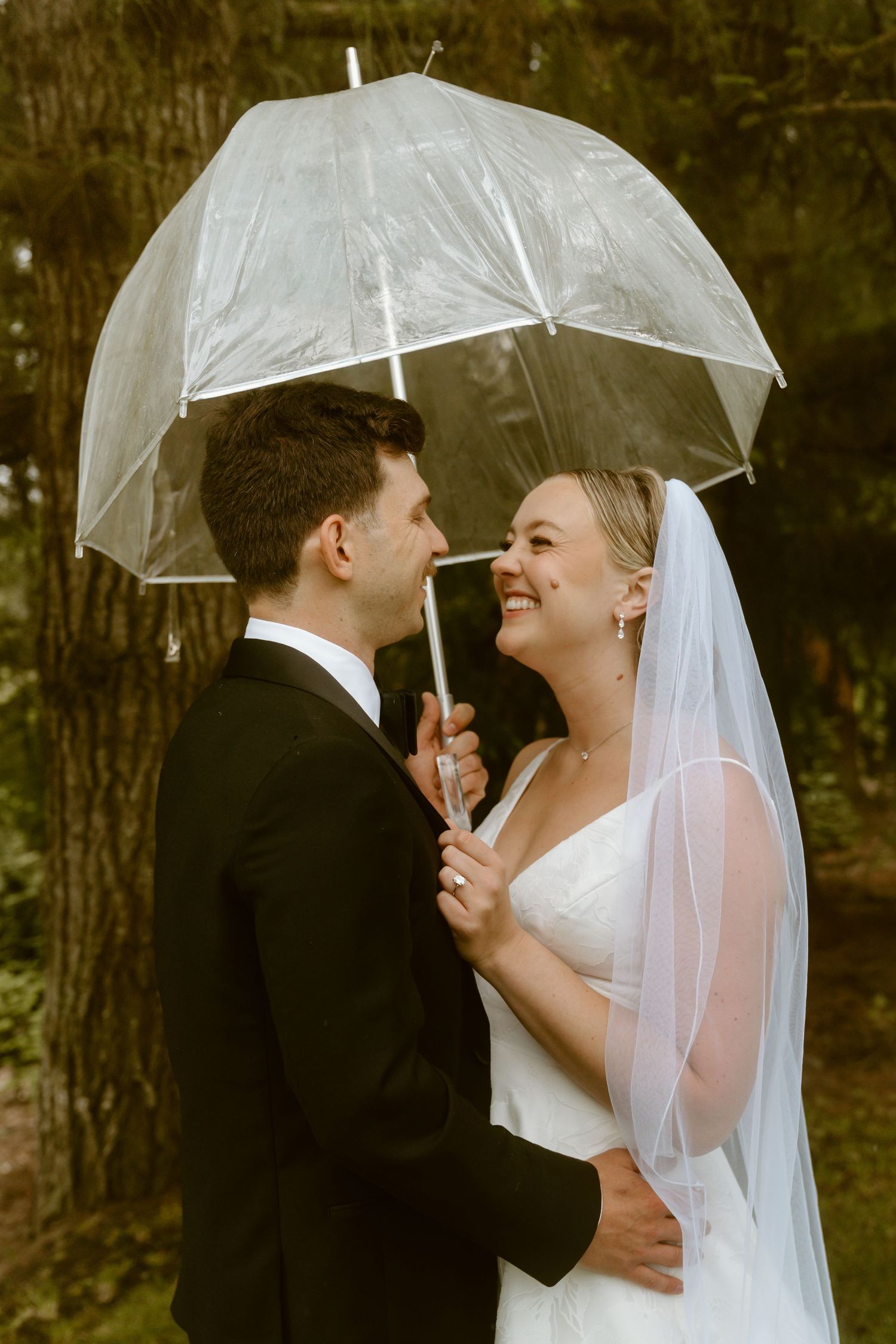 Bride and groom share a romantic moment under a clear umbrella in a lush forest setting.