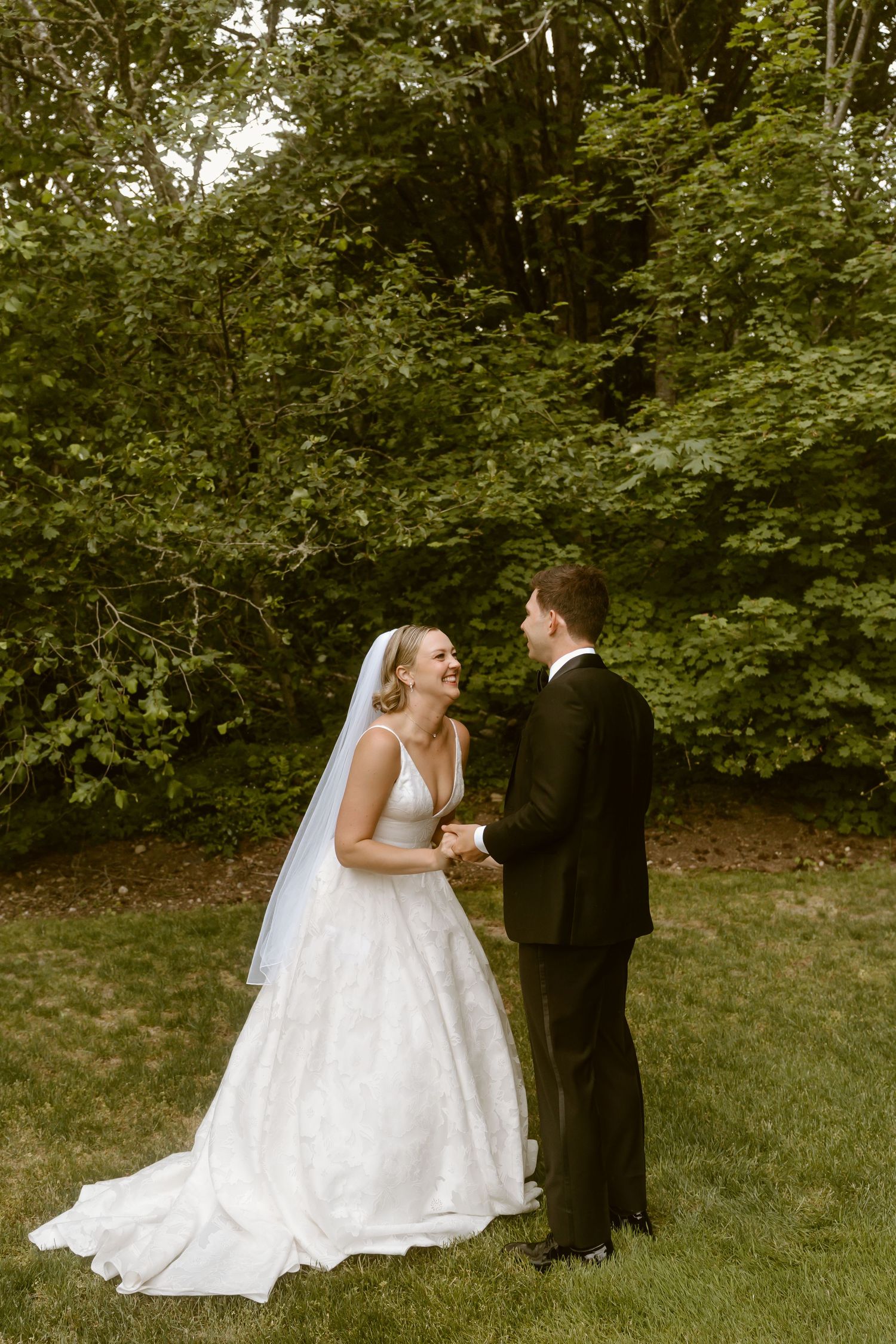 Bride and groom share a joyful first look moment on green lawn surrounded by lush trees.