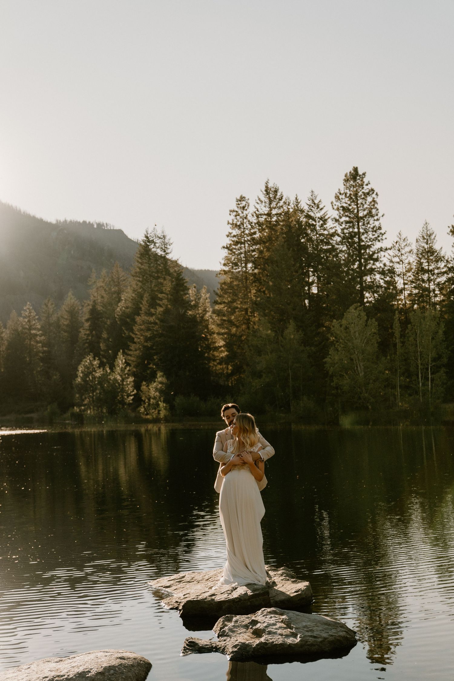 Couple in wedding attire standing on rocks by a serene mountain lake surrounded by tall pine trees at golden hour.