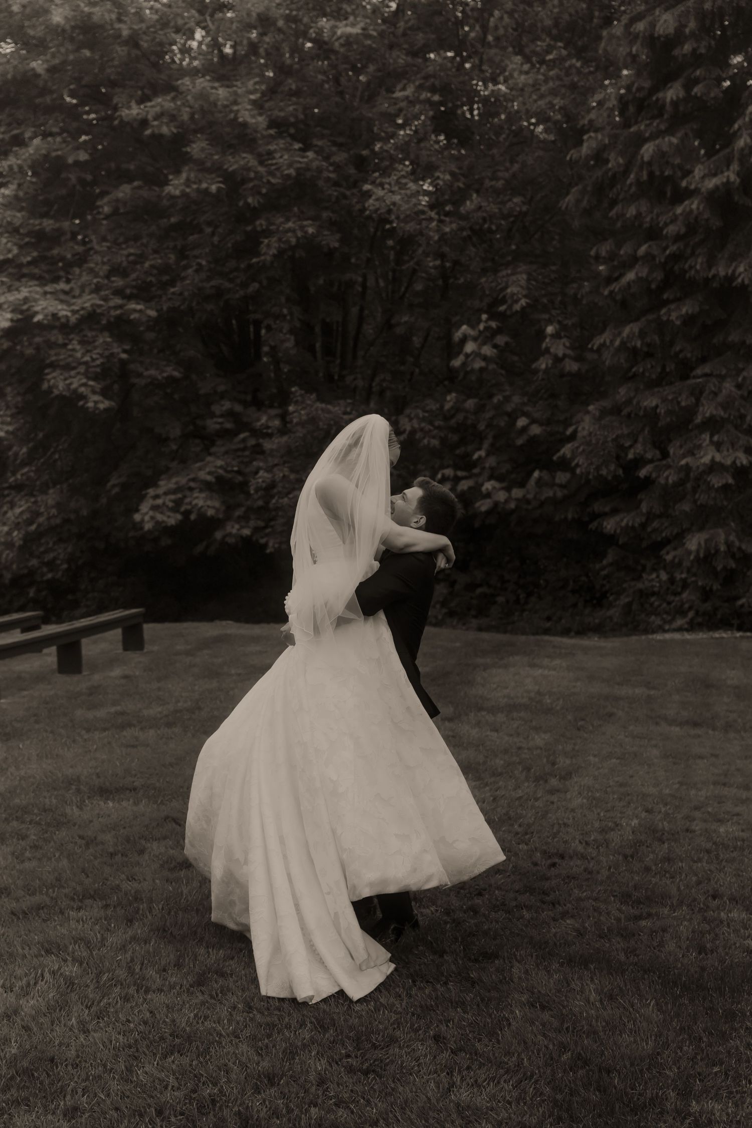 Black and white photo of groom lifting bride in joyful embrace on lush garden lawn.