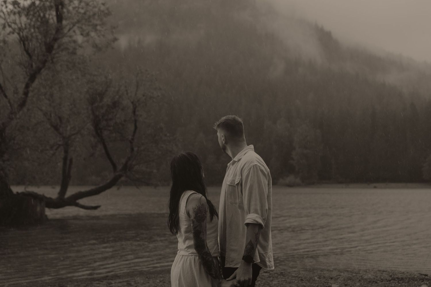 Couple holding hands facing each other in misty rain near a mountain lake in a romantic sepia-toned outdoor portrait.