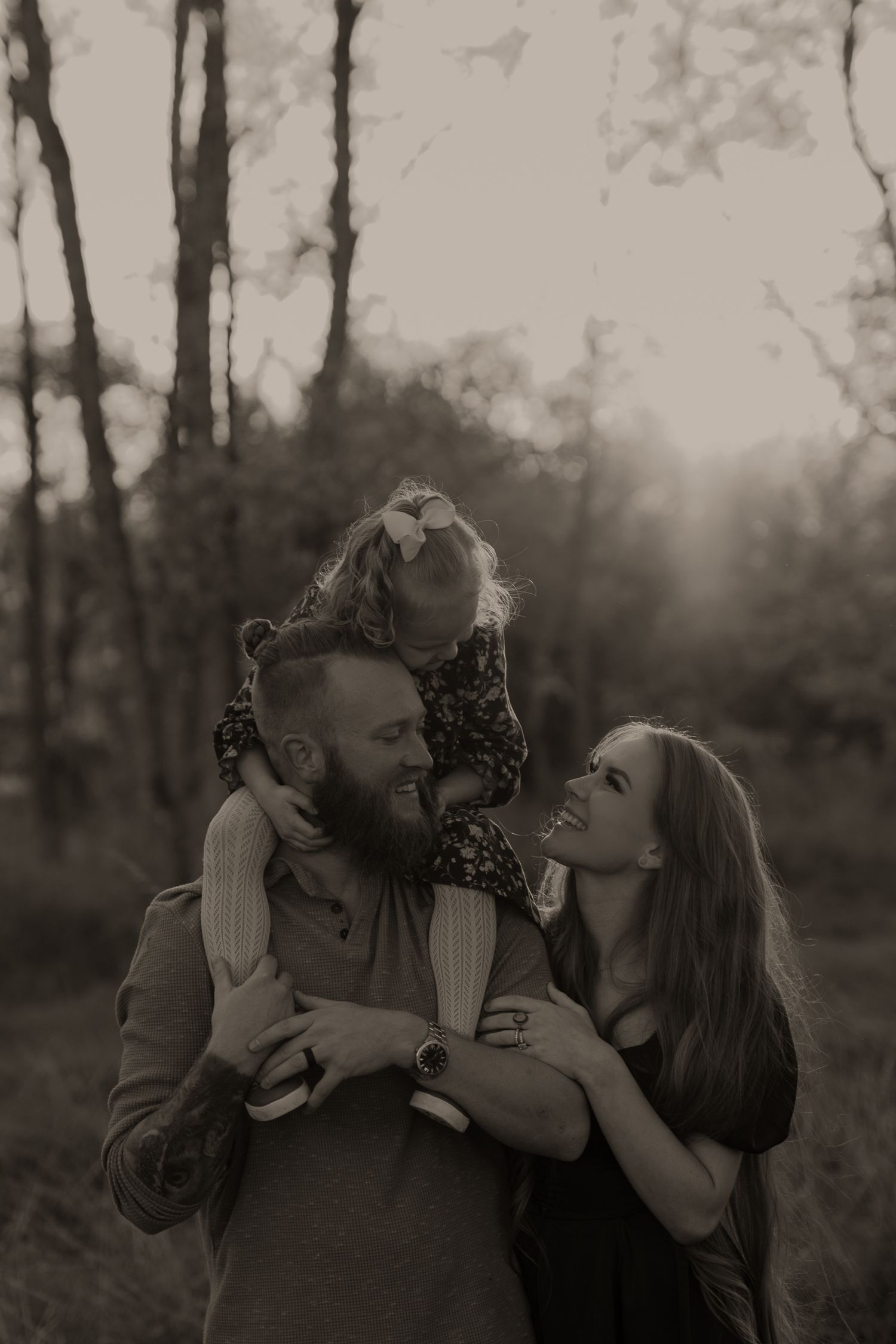 Sepia-toned family portrait of a bearded man and woman smiling at their toddler daughter perched on his shoulders outdoors.