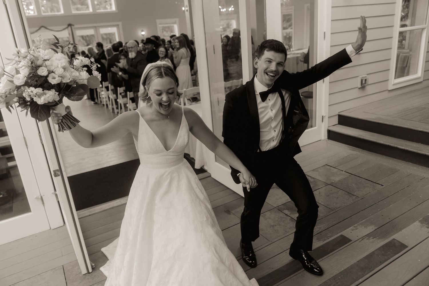 Black and white photo of joyful newlyweds dancing at wedding reception entrance, bride in gown holding bouquet.