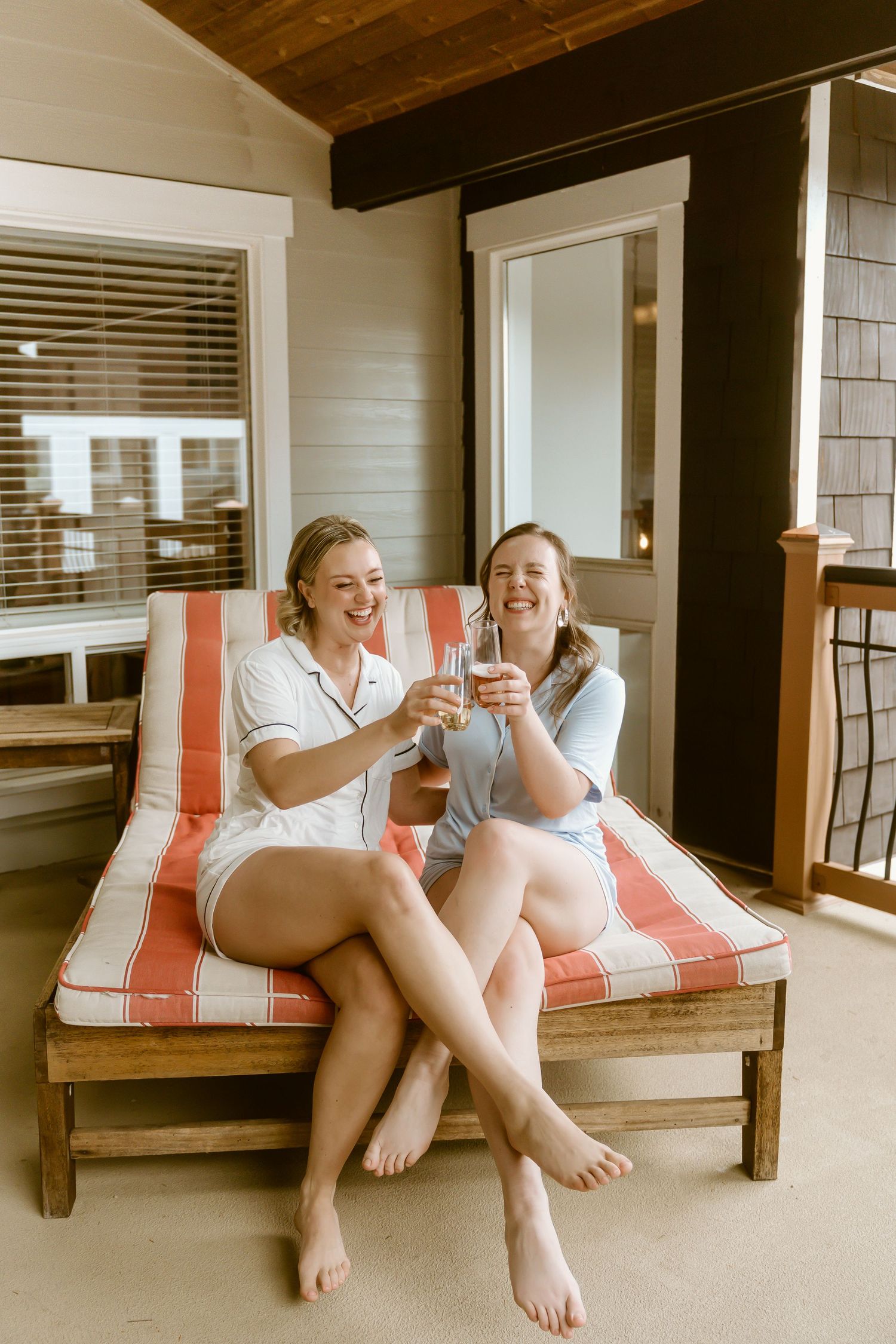 Two women laughing and toasting drinks on a striped lounge chair on a cozy porch.
