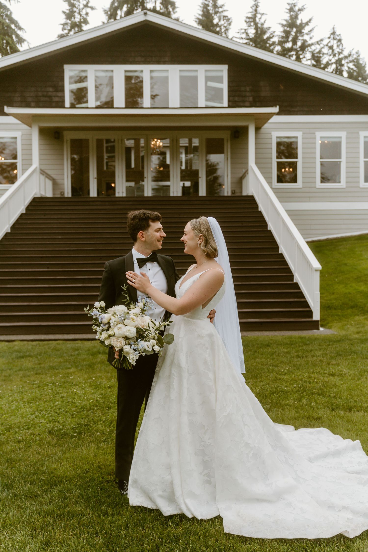 Bride and groom share a joyful moment on green lawn before elegant gray wedding venue with grand staircase.