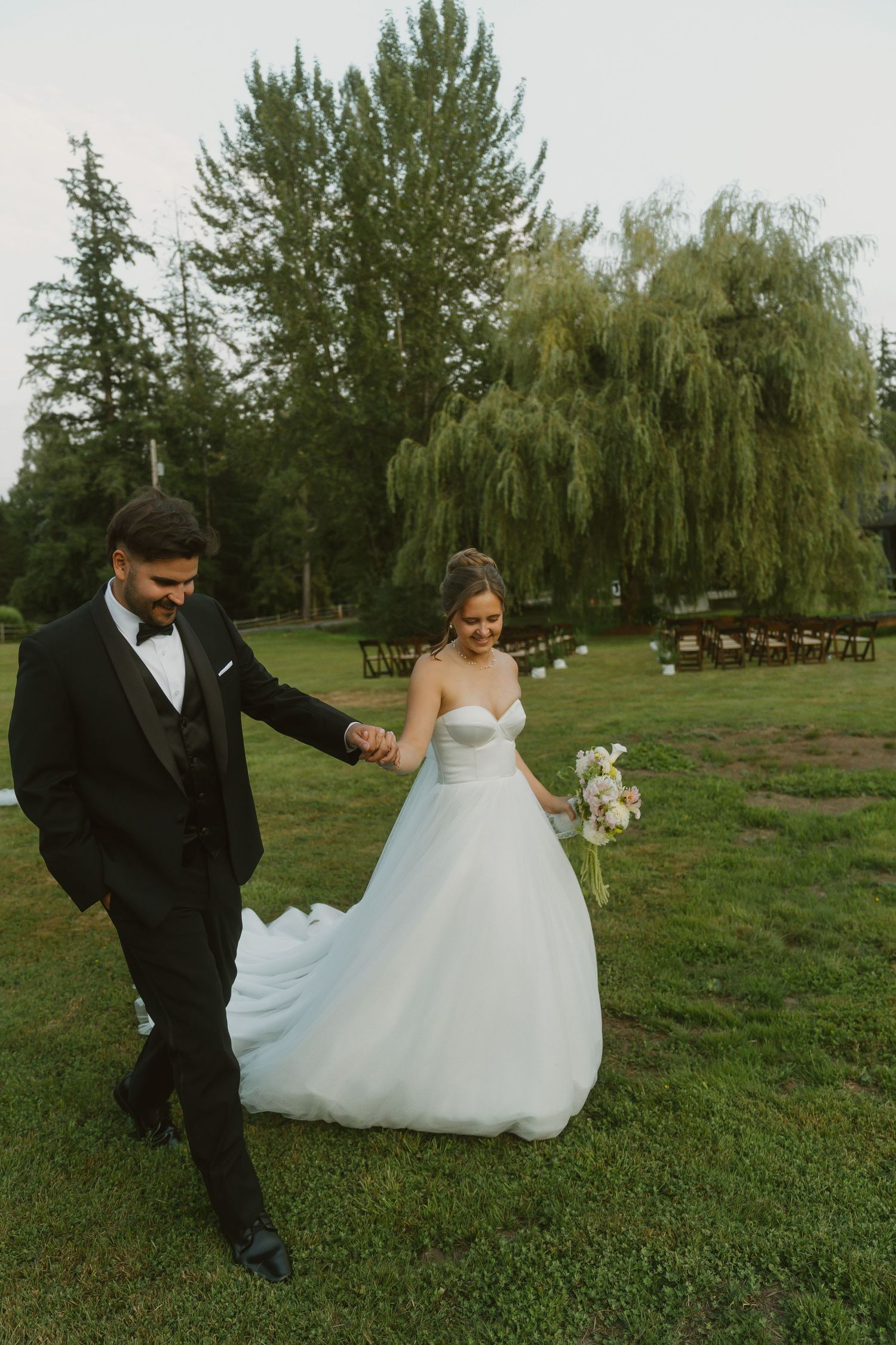 Bride in a strapless white ball gown holding a bouquet walks hand in hand with a groom in a black tuxedo across a grassy outdoor wedding ven
