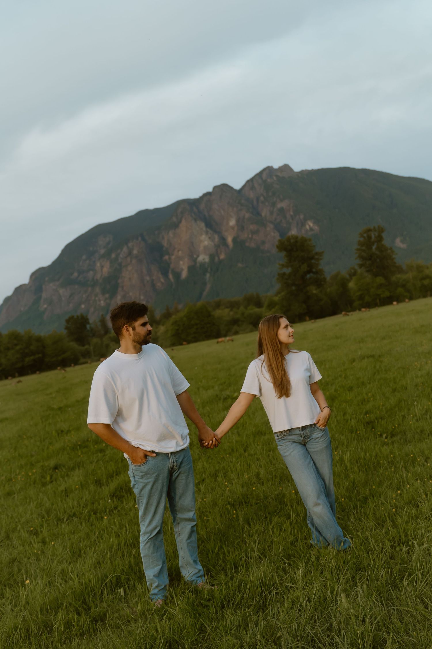 Couple in matching white t-shirts and jeans holding hands in a green field with a mountain backdrop.