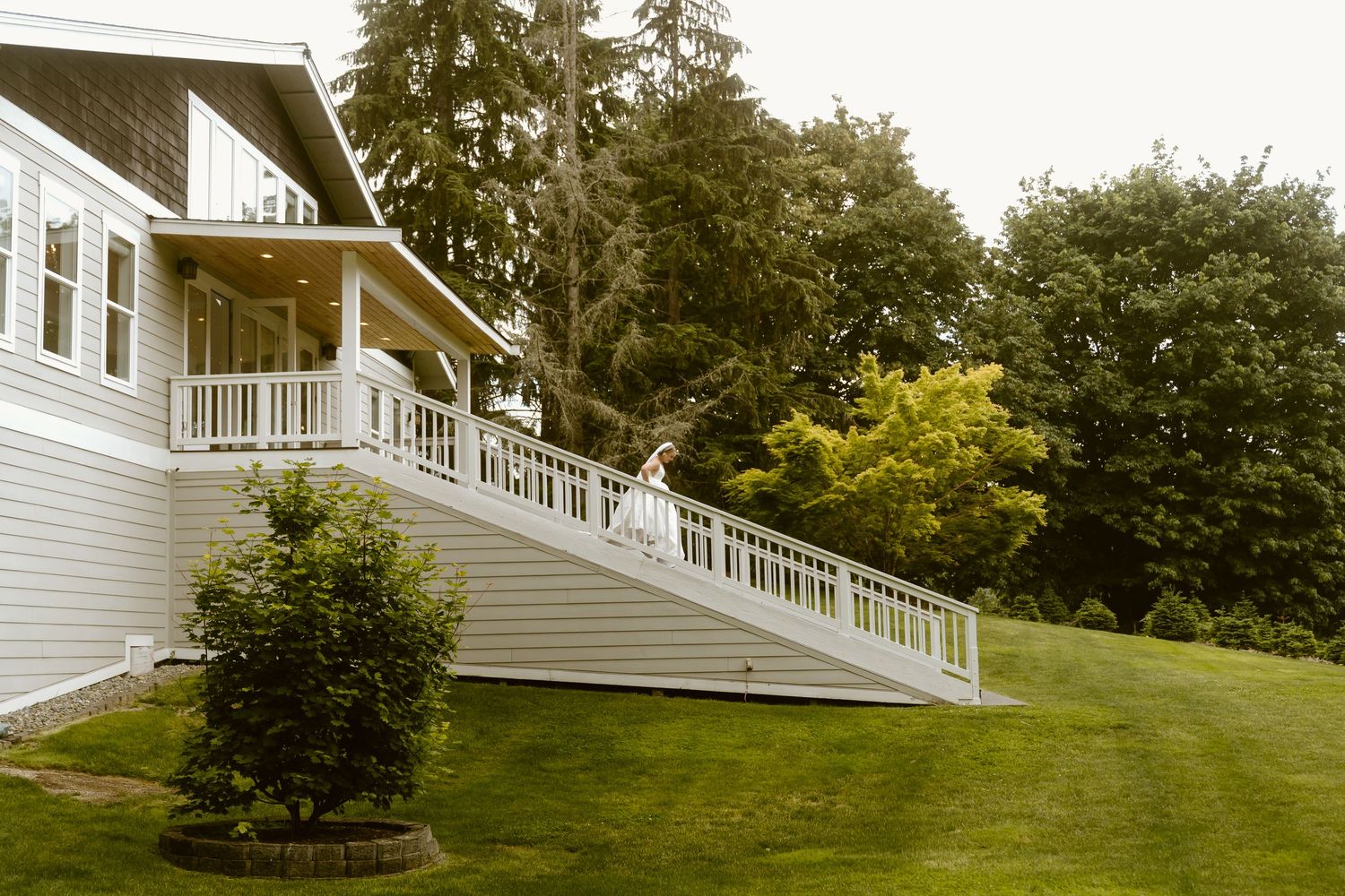 Bride standing on white railed deck of elegant gray house surrounded by lush green trees.