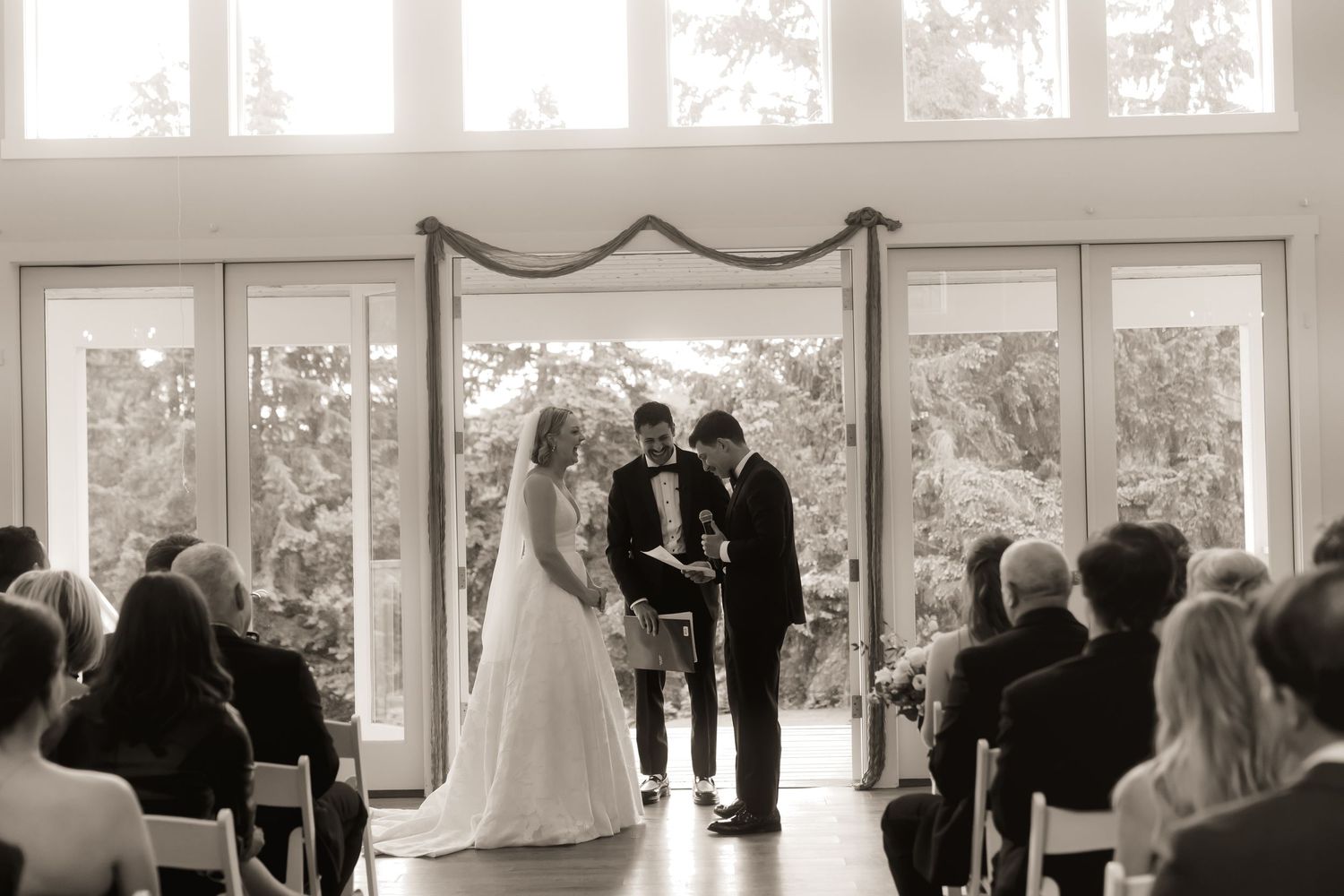 Black and white photo of a couple exchanging vows at an indoor wedding ceremony with large windows and seated guests.