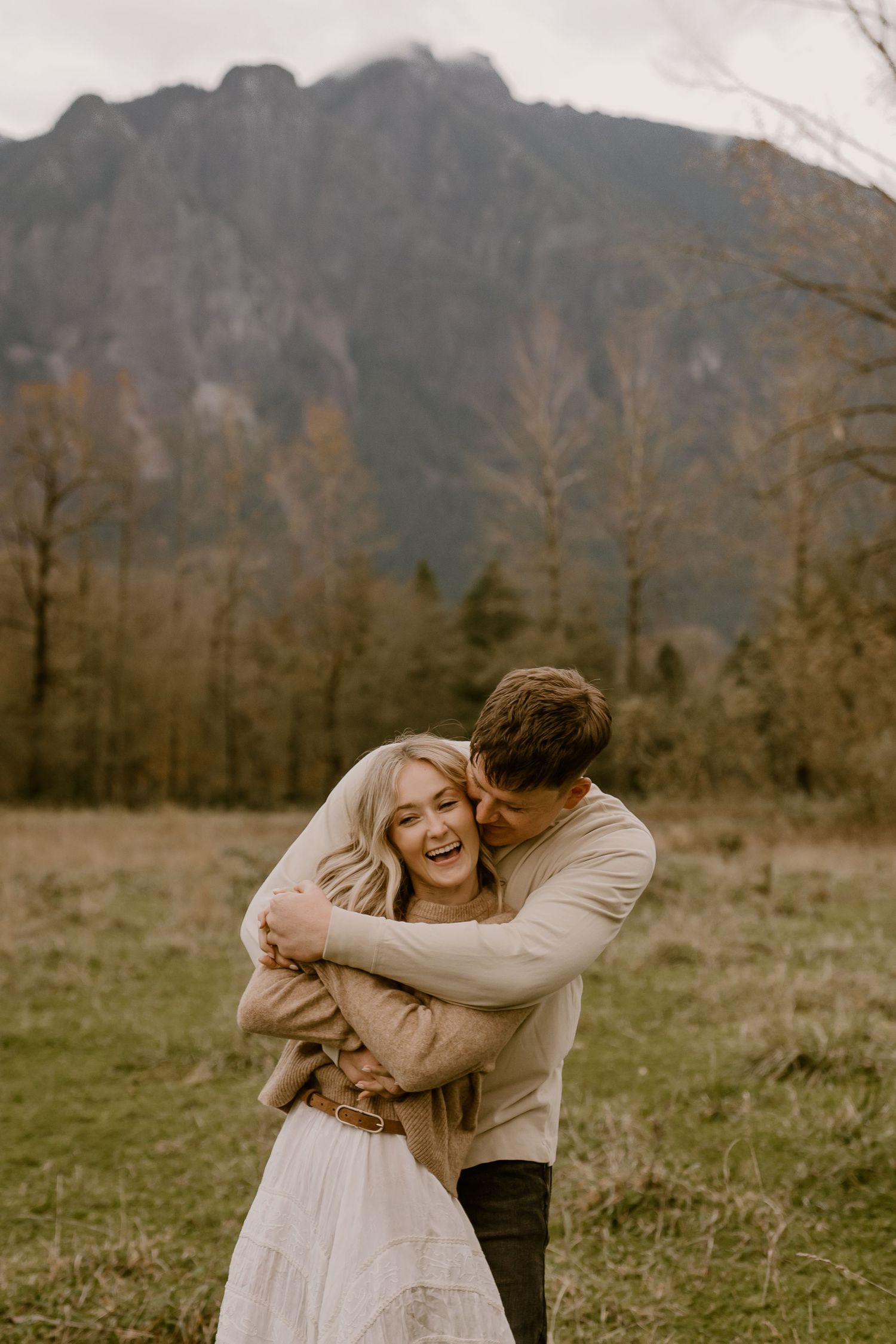 A couple embraces and laughs in a mountain meadow, the man hugging the woman from behind during their engagement session.