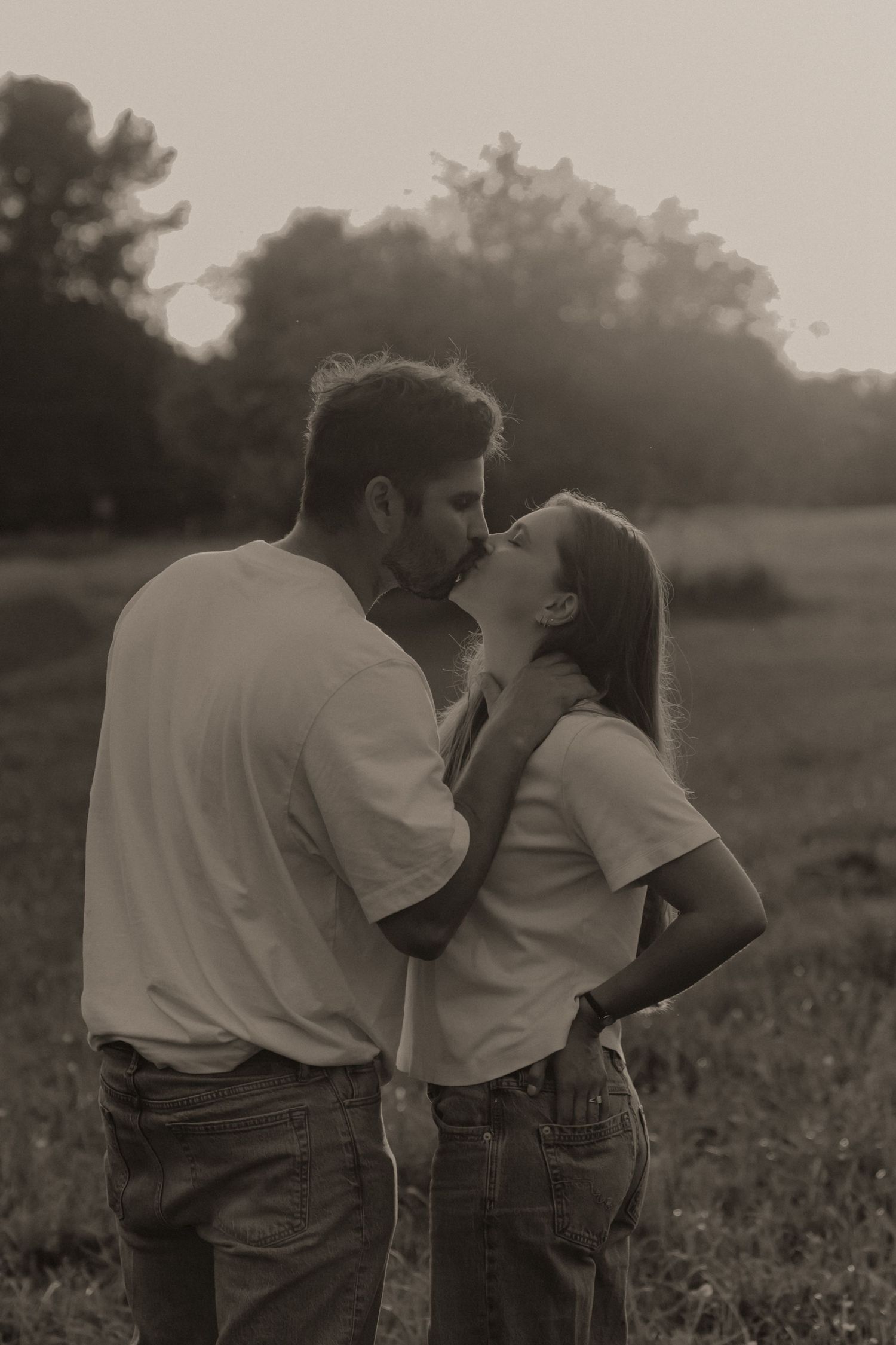 Romantic sepia-toned photo of a couple sharing a tender kiss in an outdoor field at dusk.