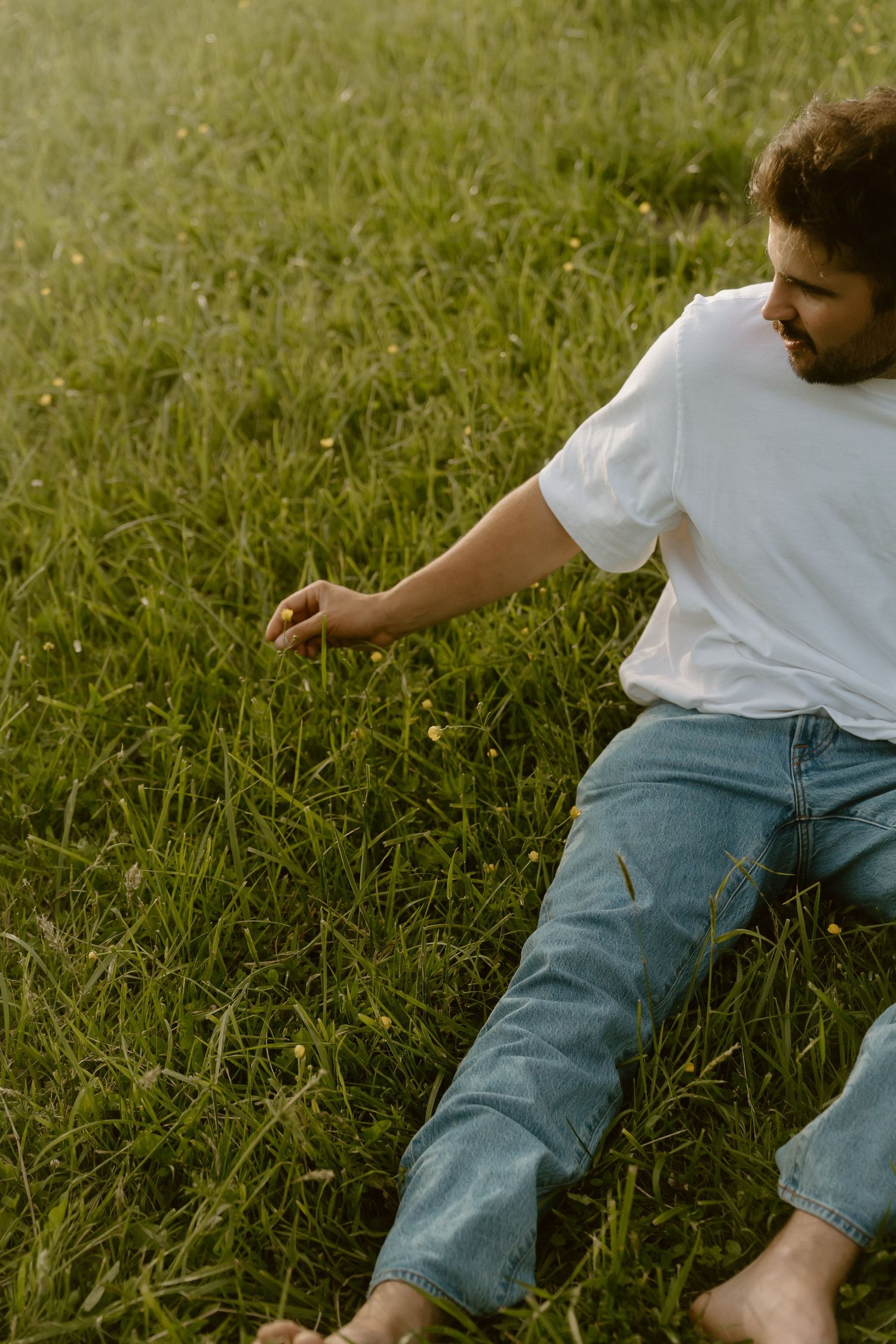 Man in white t-shirt and jeans sitting barefoot on lush green grass, reaching down to touch wildflowers.