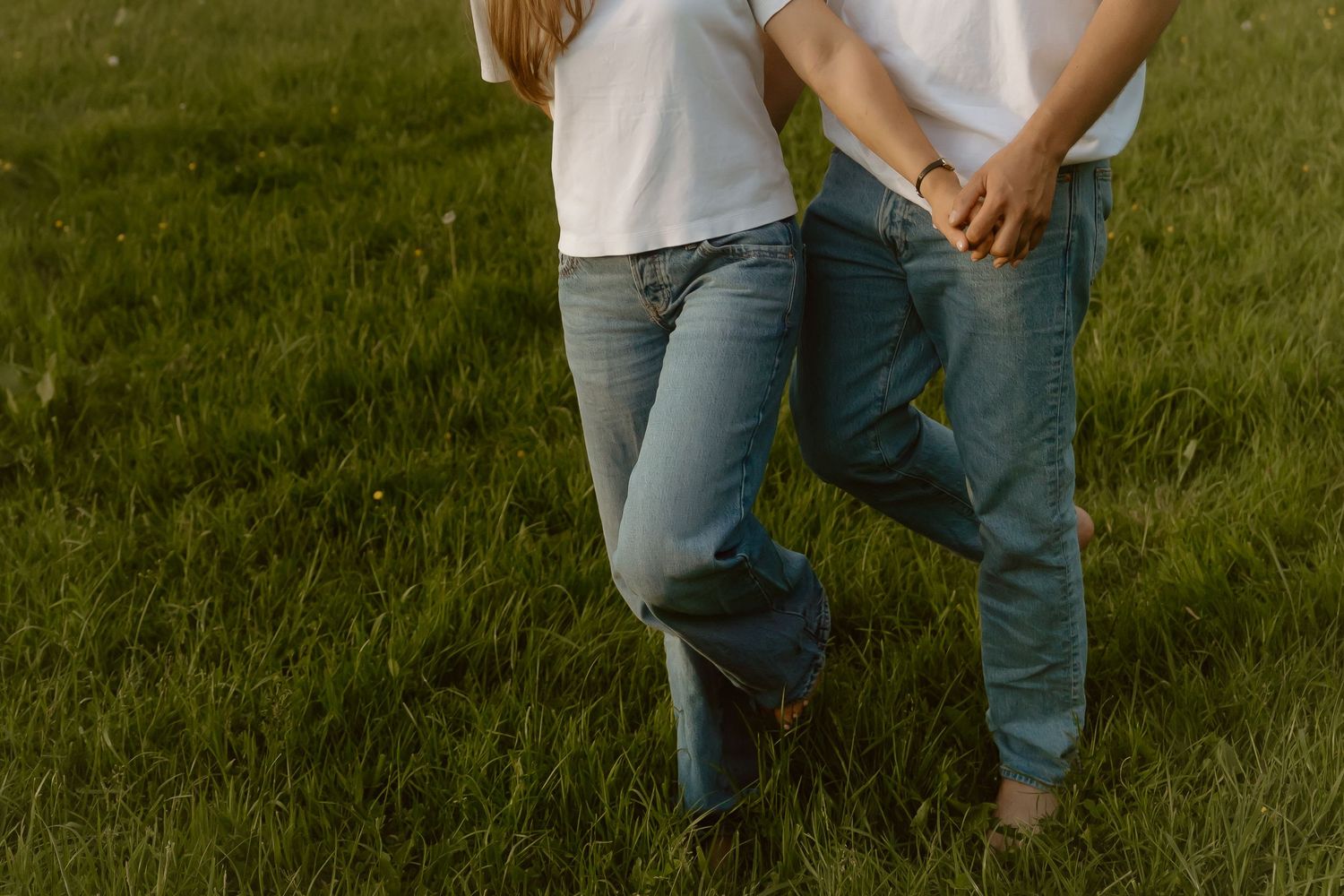 Couple holding hands in green field wearing white shirts and blue jeans, romantic outdoor lifestyle photo.
