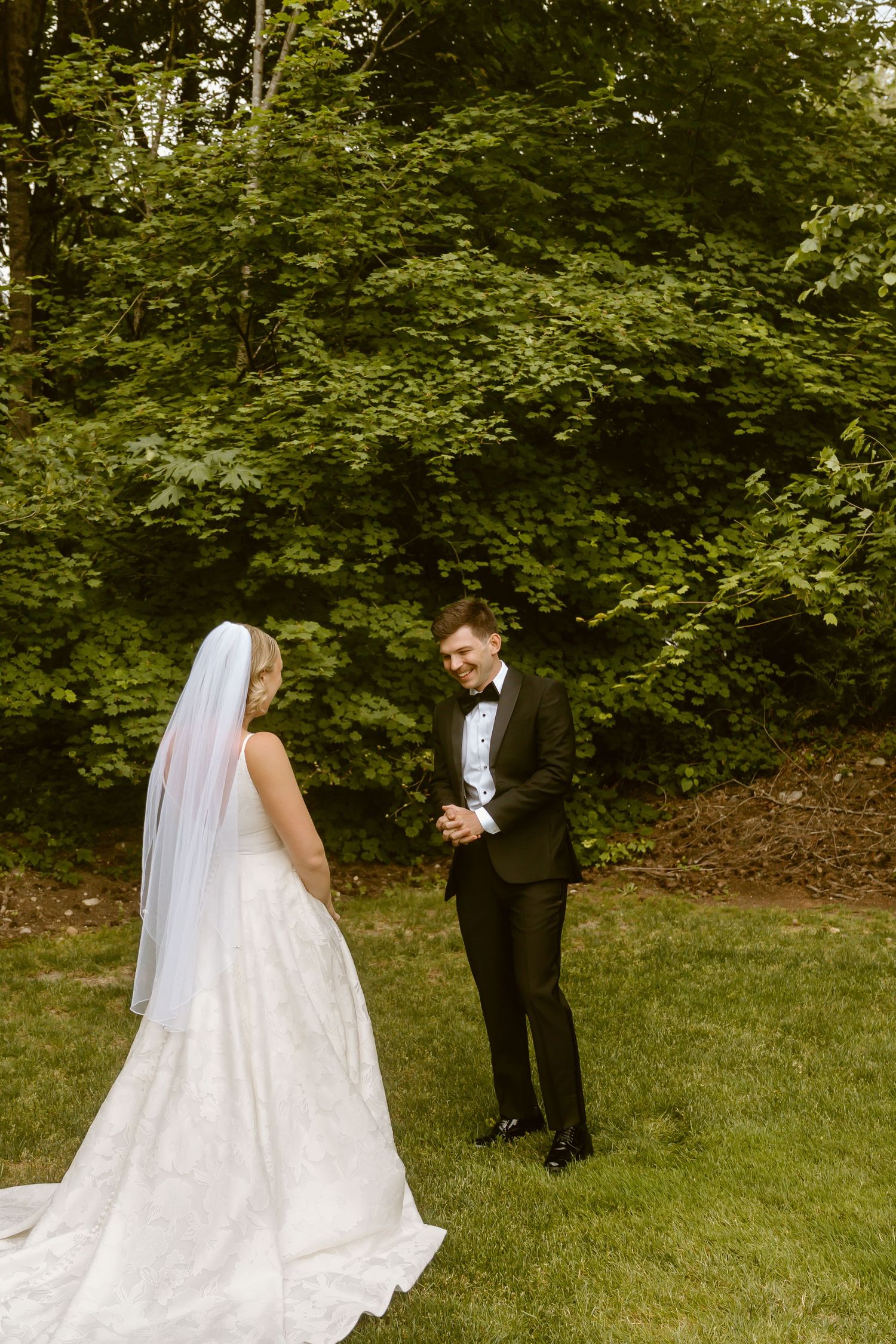 Groom smiling at bride during outdoor first look surrounded by lush green trees.