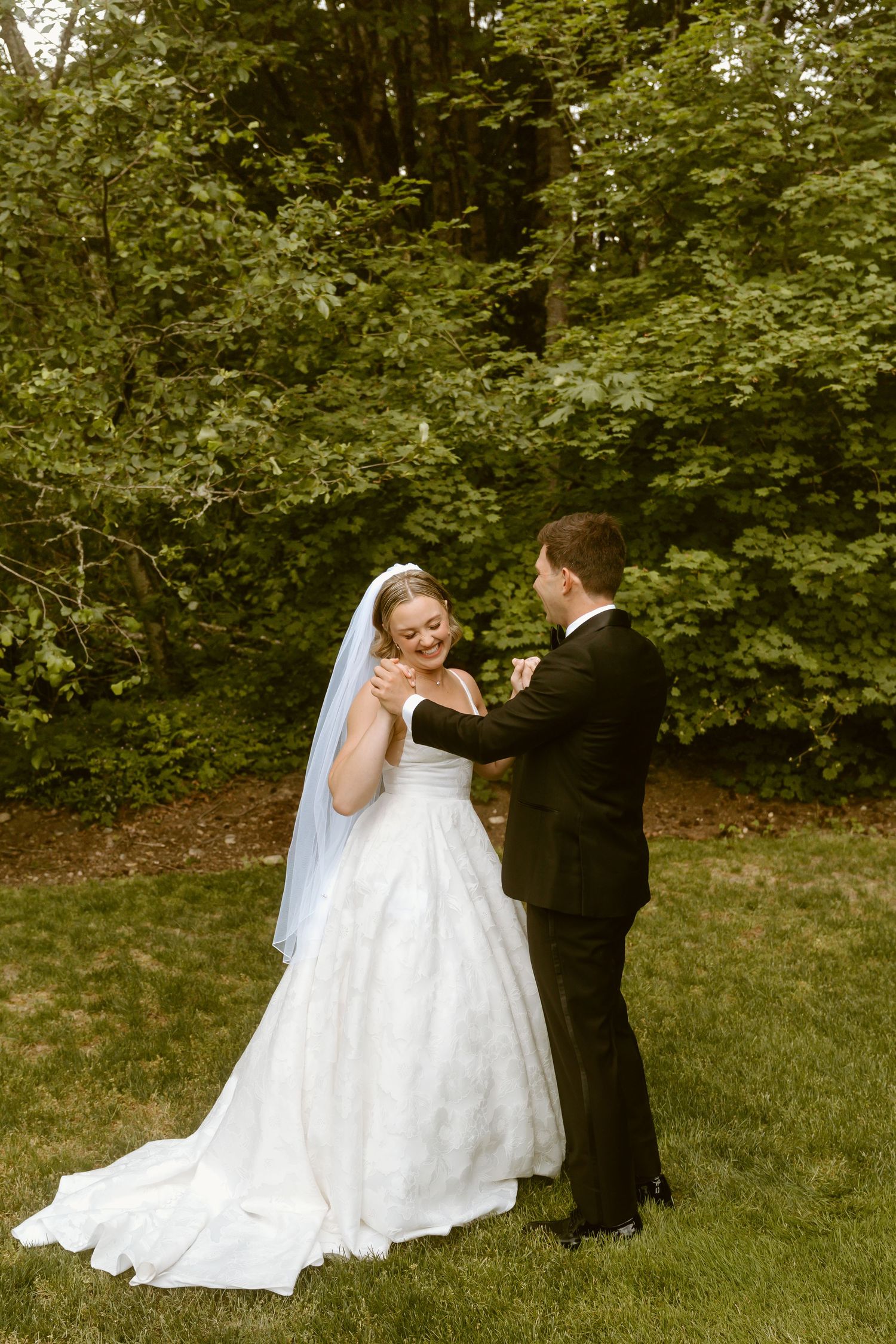 Bride and groom share a joyful first look moment on a lush green lawn surrounded by tall trees.
