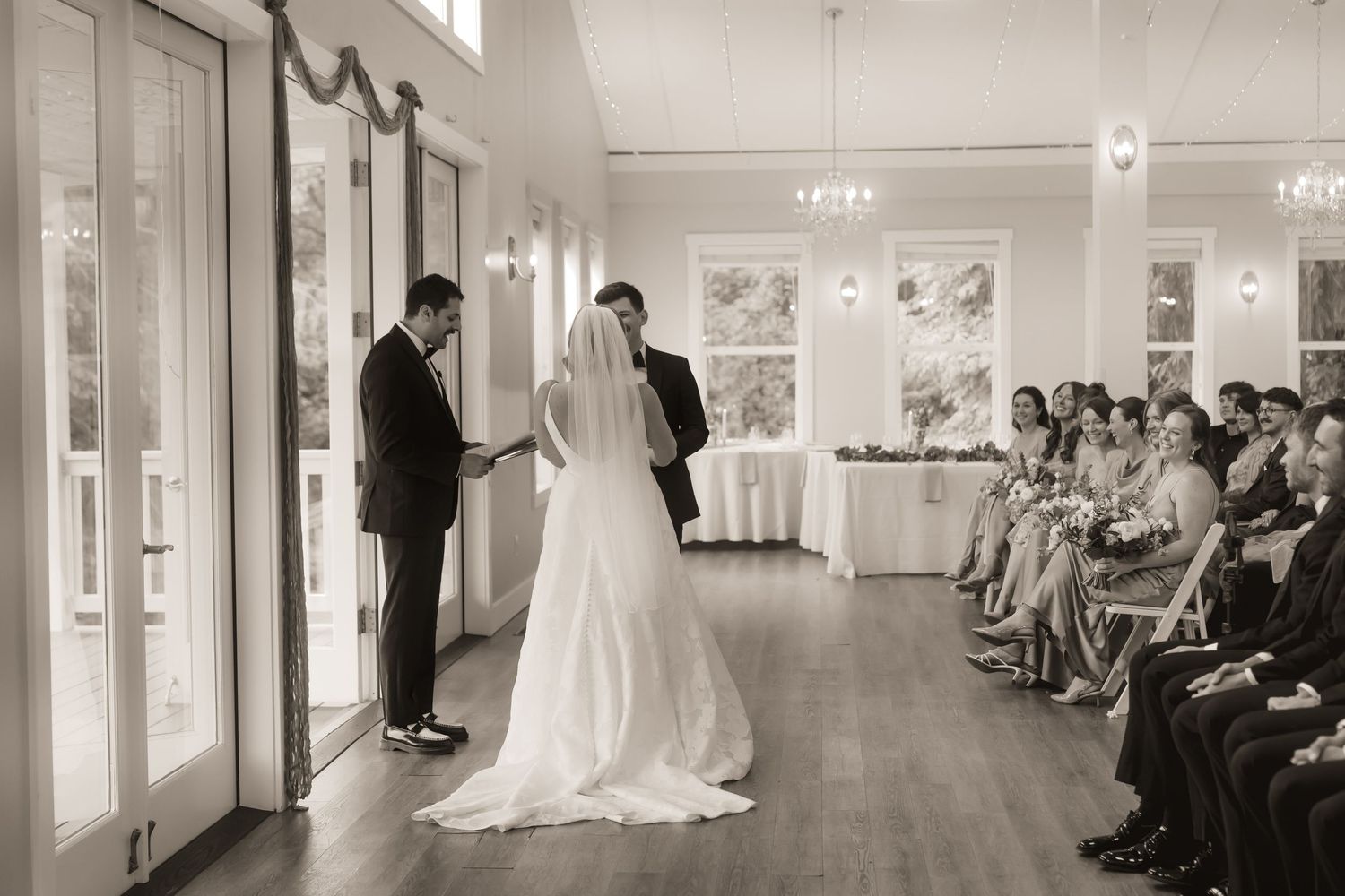 Black and white photo of a bride and groom exchanging vows at an elegant indoor wedding ceremony with guests seated.