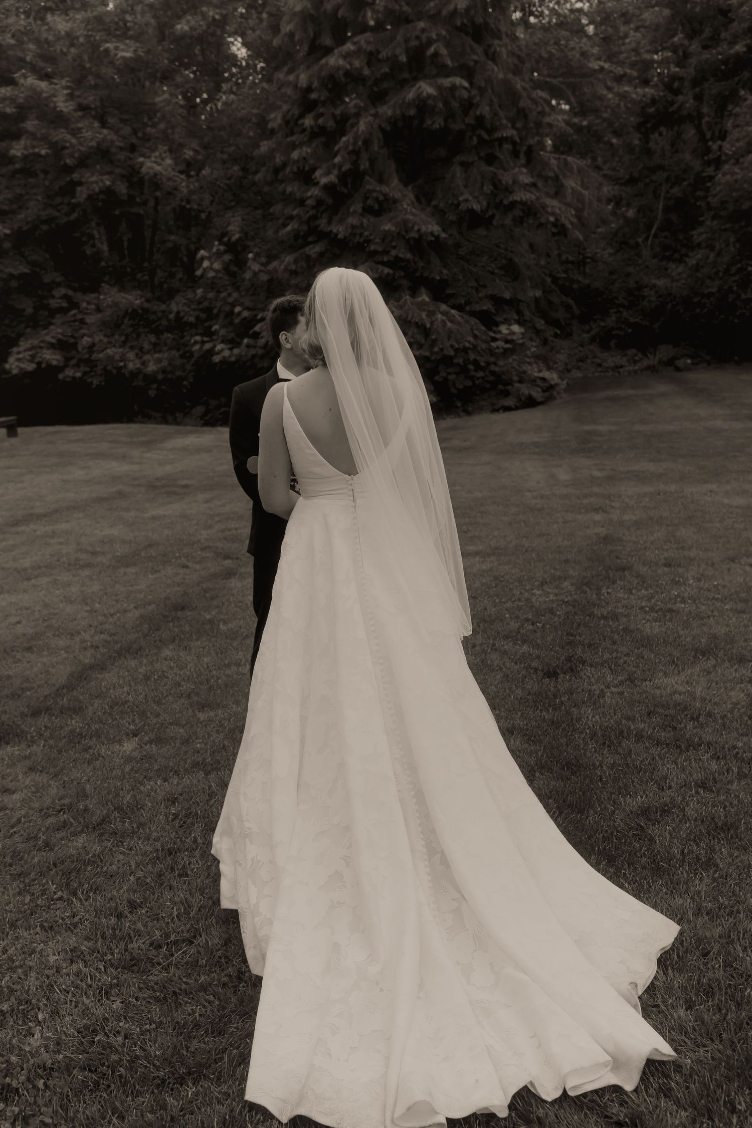 Black and white photo of bride in flowing white gown and veil walking with groom on lawn.