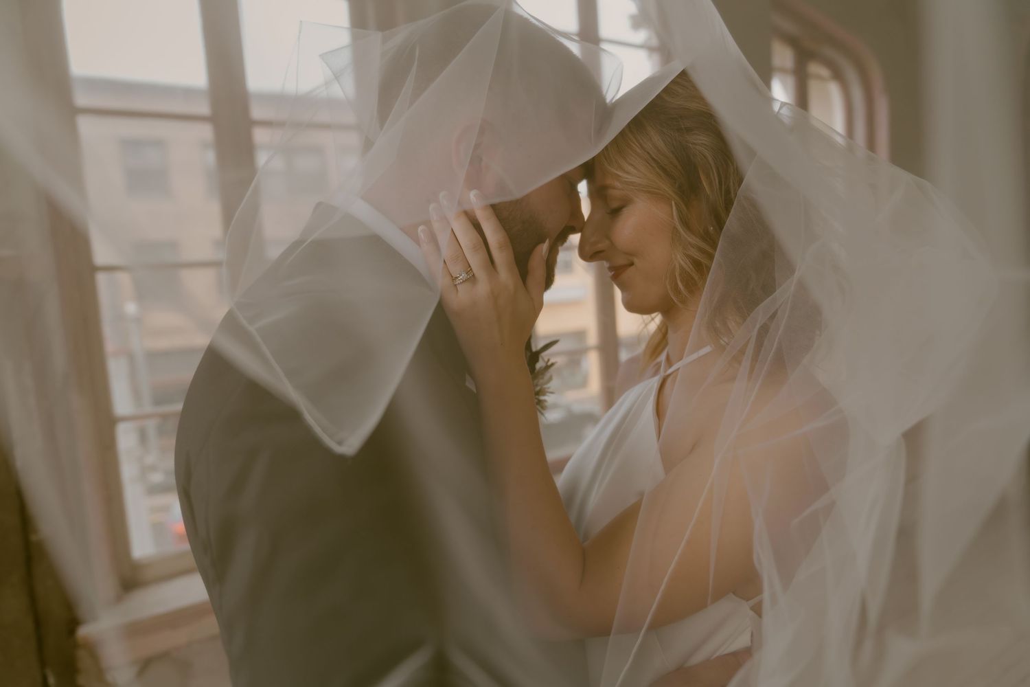 A romantic wedding moment captured through soft sheer veil in warm vintage lighting by a window.