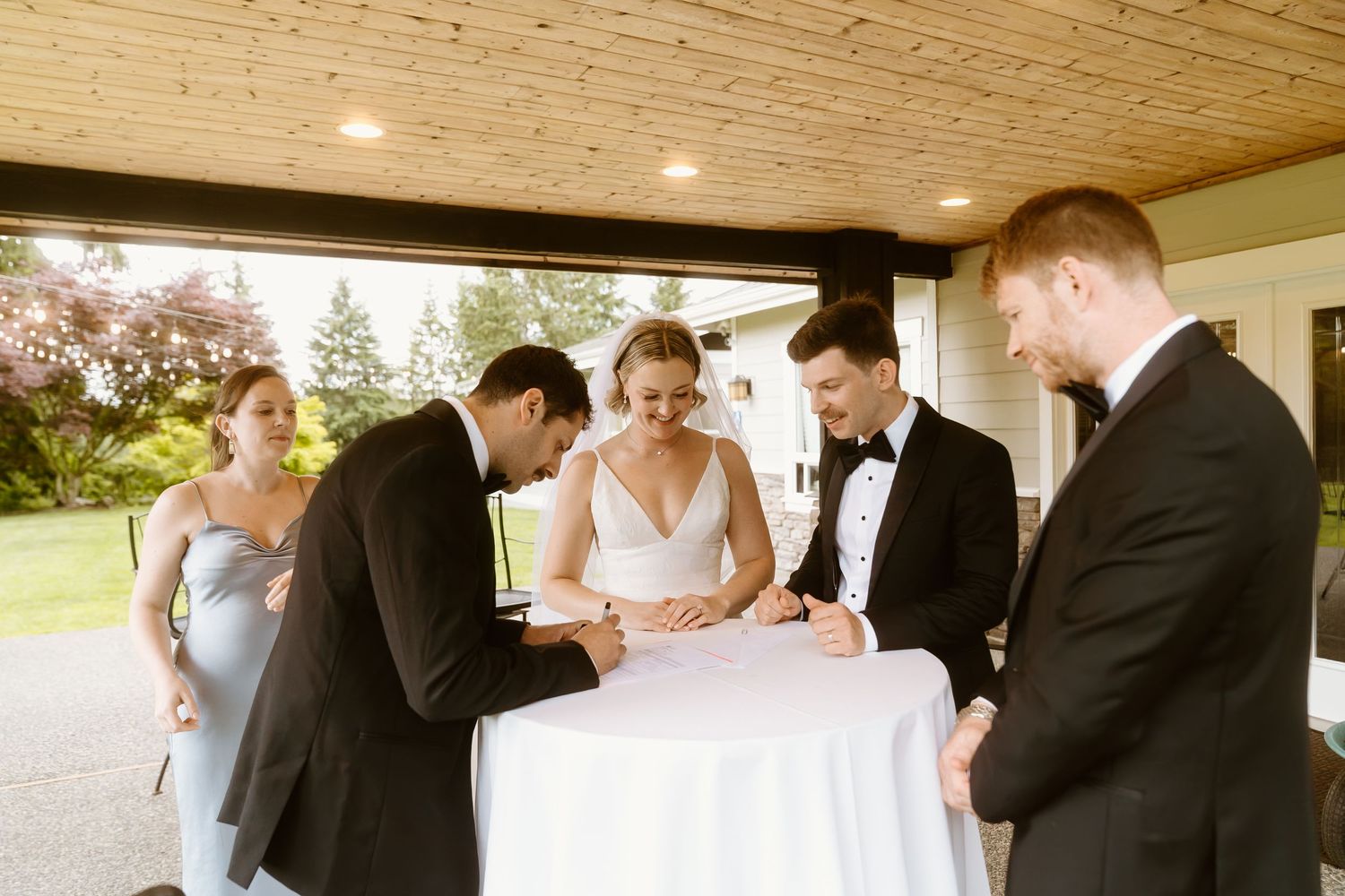 Wedding party signs marriage documents at outdoor table with bride and groom.