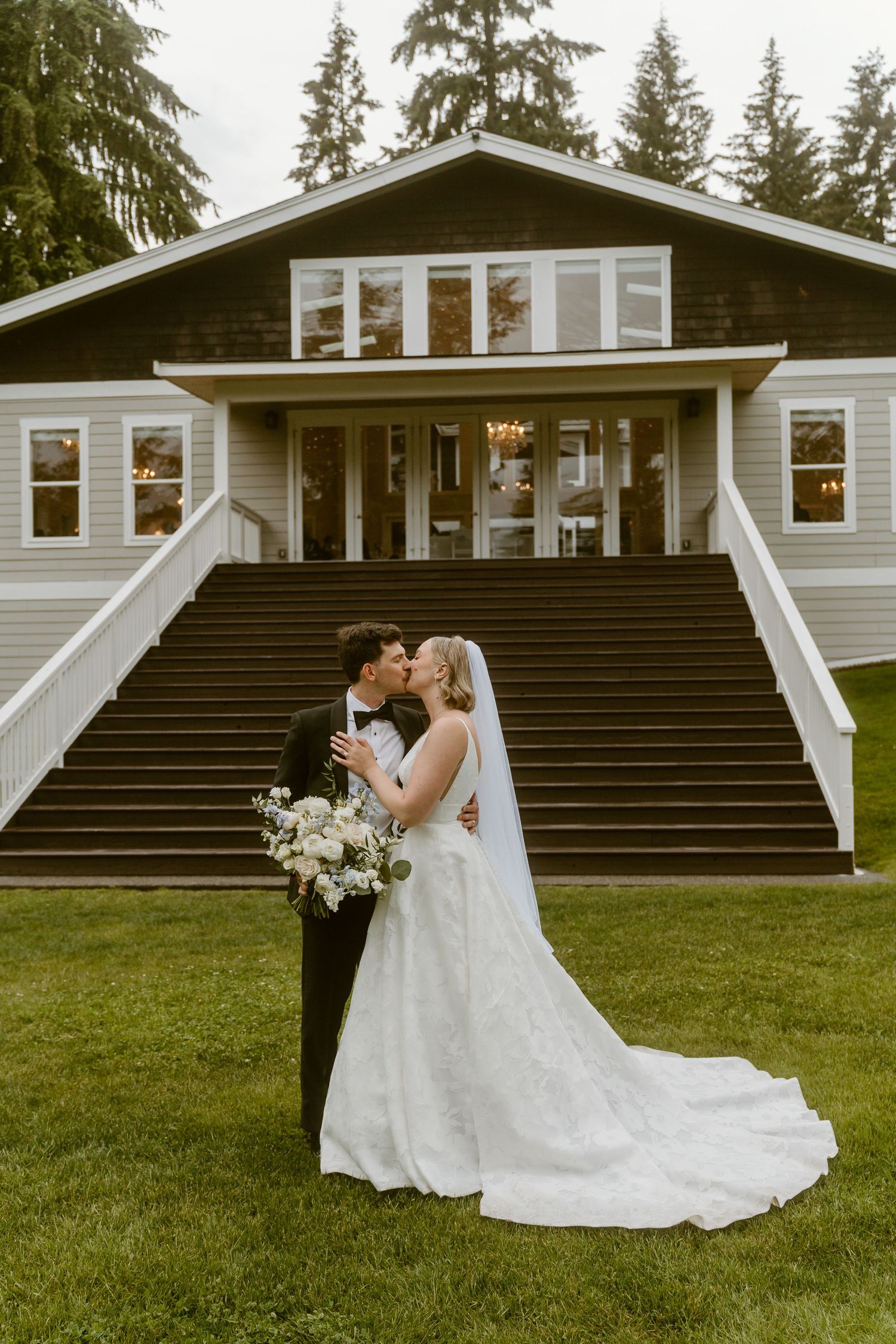 Bride and groom kissing in front of elegant venue staircase, bride holding white floral bouquet in wedding gown.