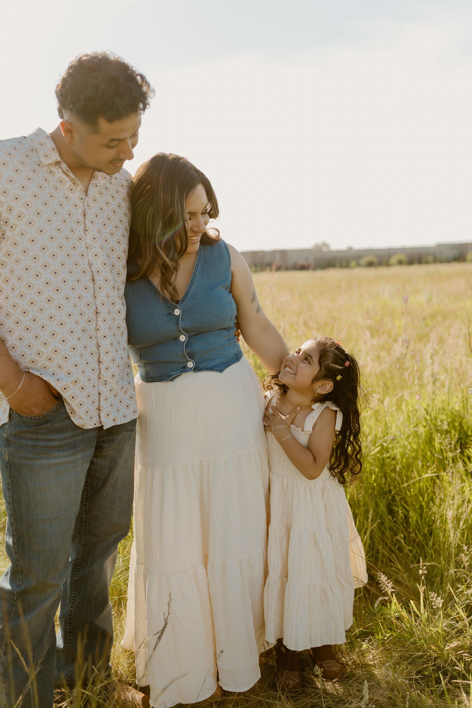 Family of three posing in a sunlit field, parents smiling at young daughter in matching cream dresses.