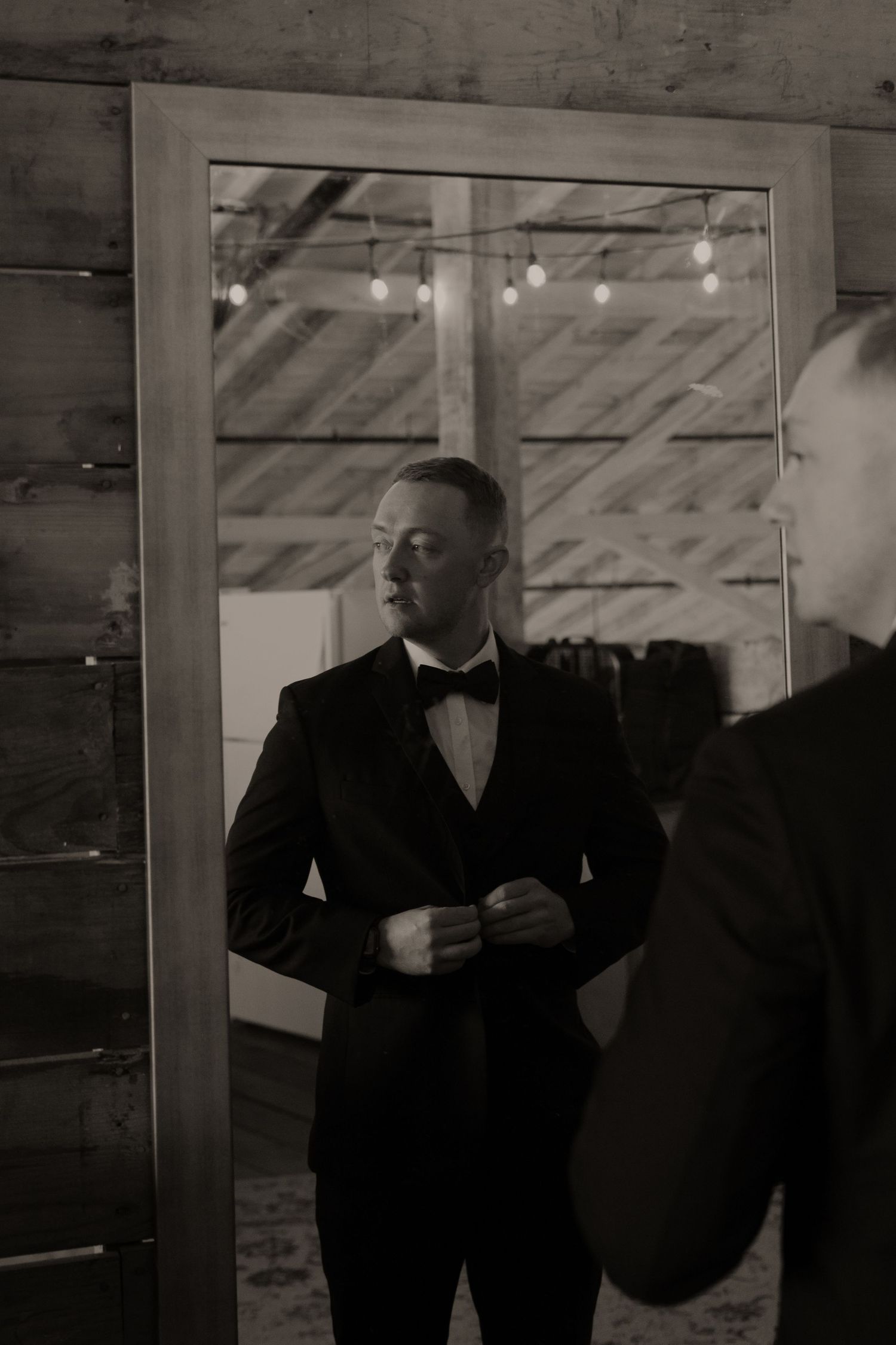 Groom adjusting suit in mirror reflection at rustic wooden venue.