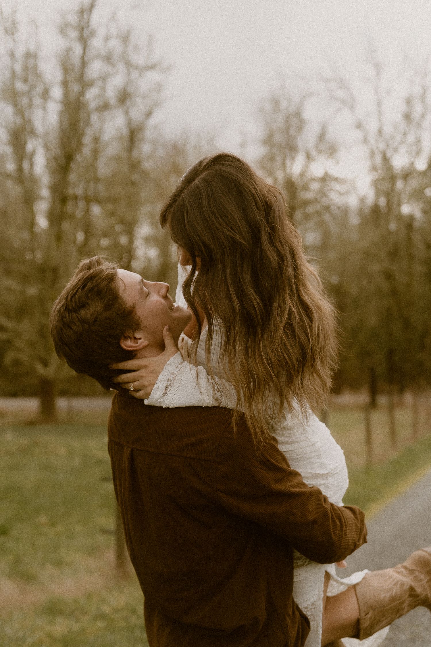 A man lifts a woman in a romantic embrace outdoors, surrounded by bare trees in a soft, muted landscape.