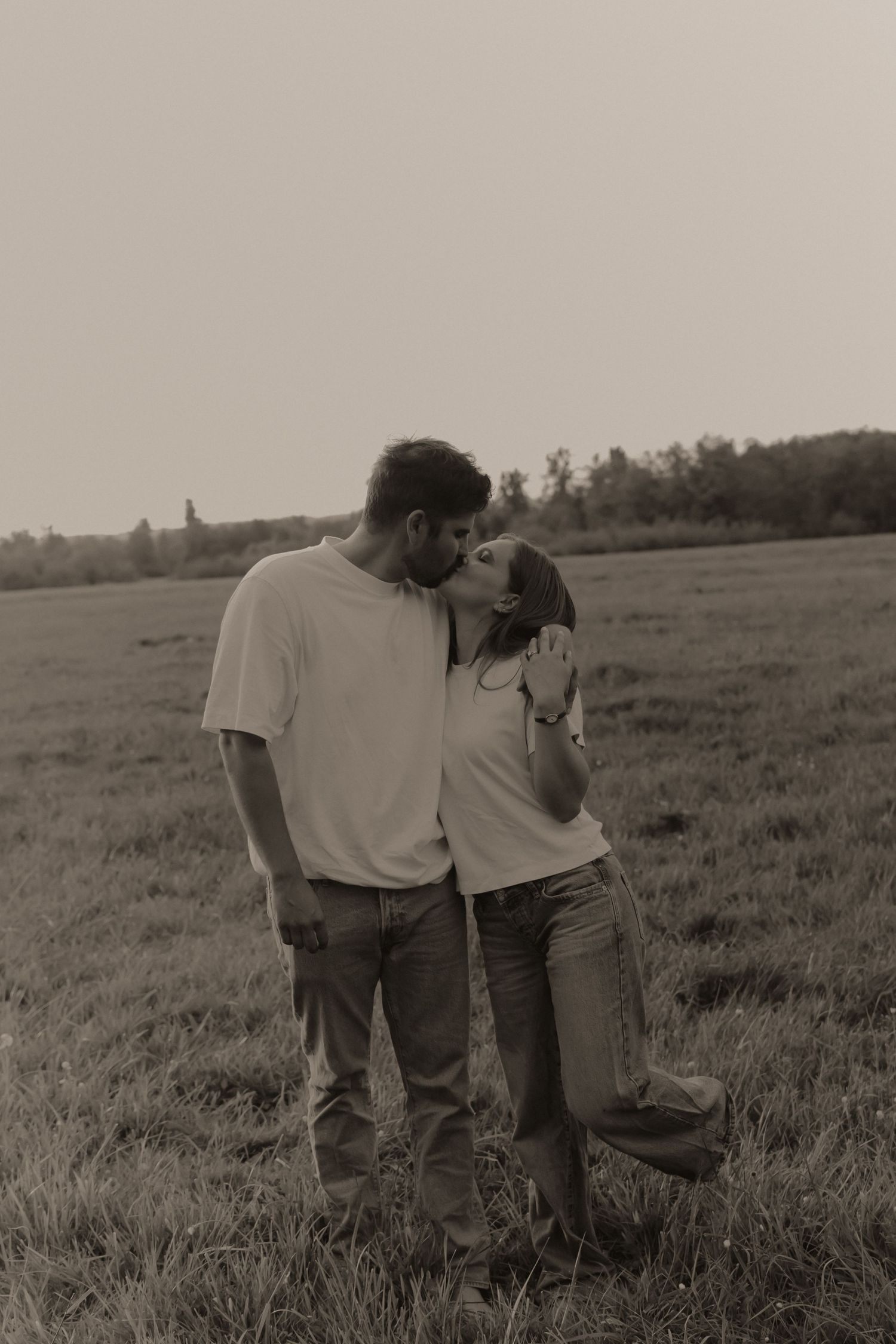Couple sharing a tender kiss in a sepia-toned field, wearing white shirts and jeans in a romantic outdoor setting.