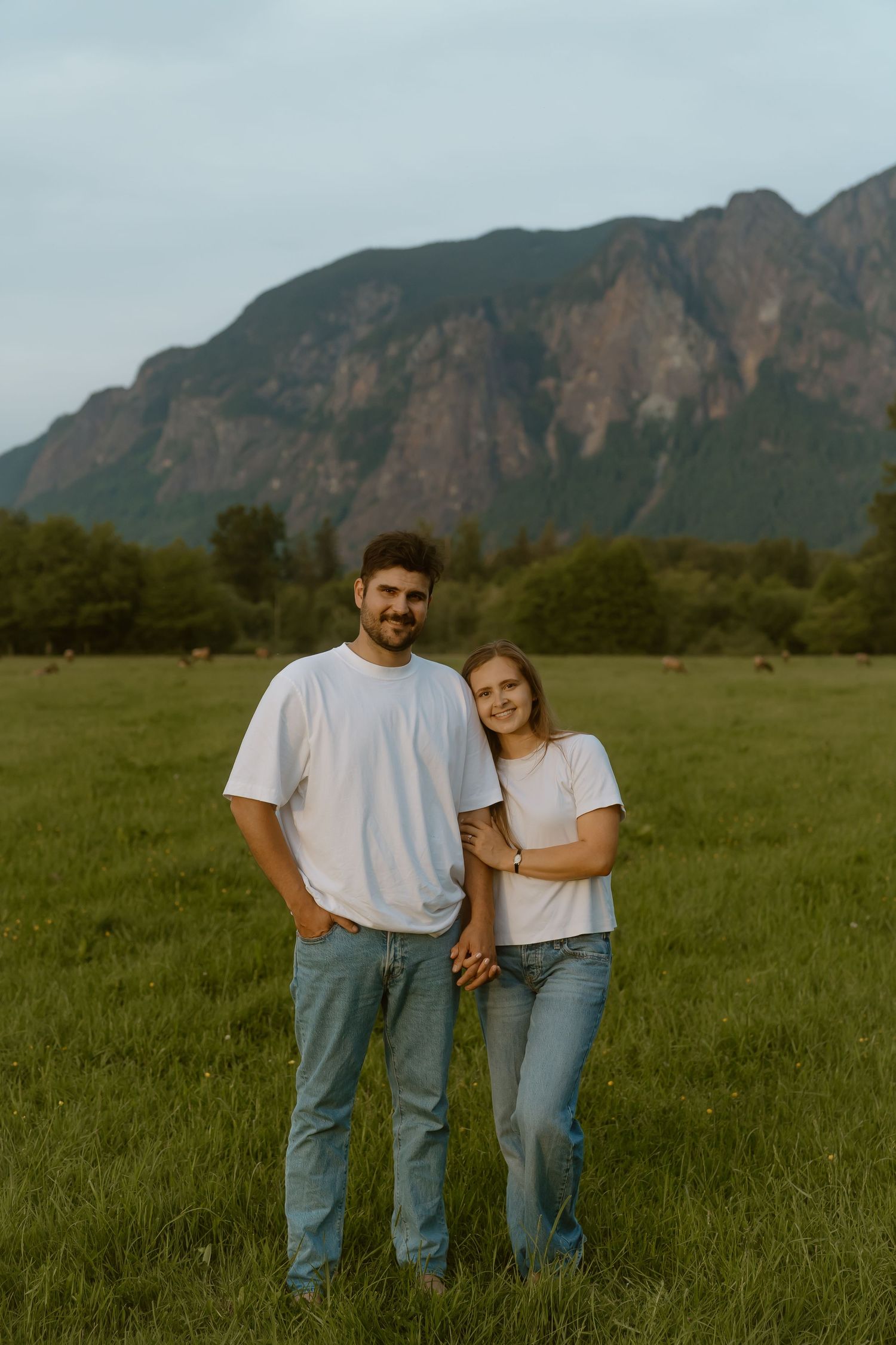 Couple in matching white t-shirts and jeans holding hands in a green field with a mountain backdrop.