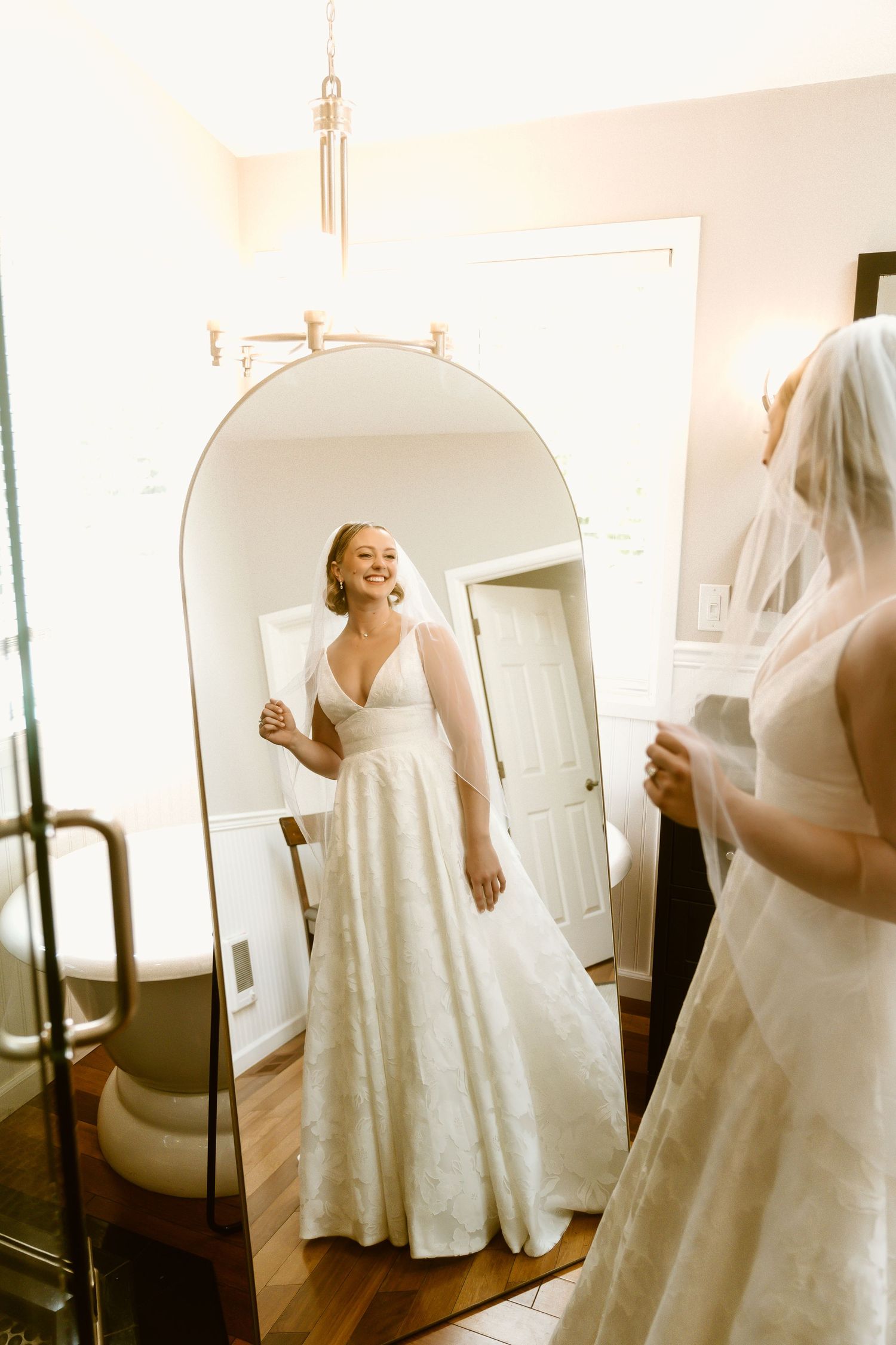 A smiling bride in a white gown and veil admires her reflection in an arched floor mirror on her wedding day.