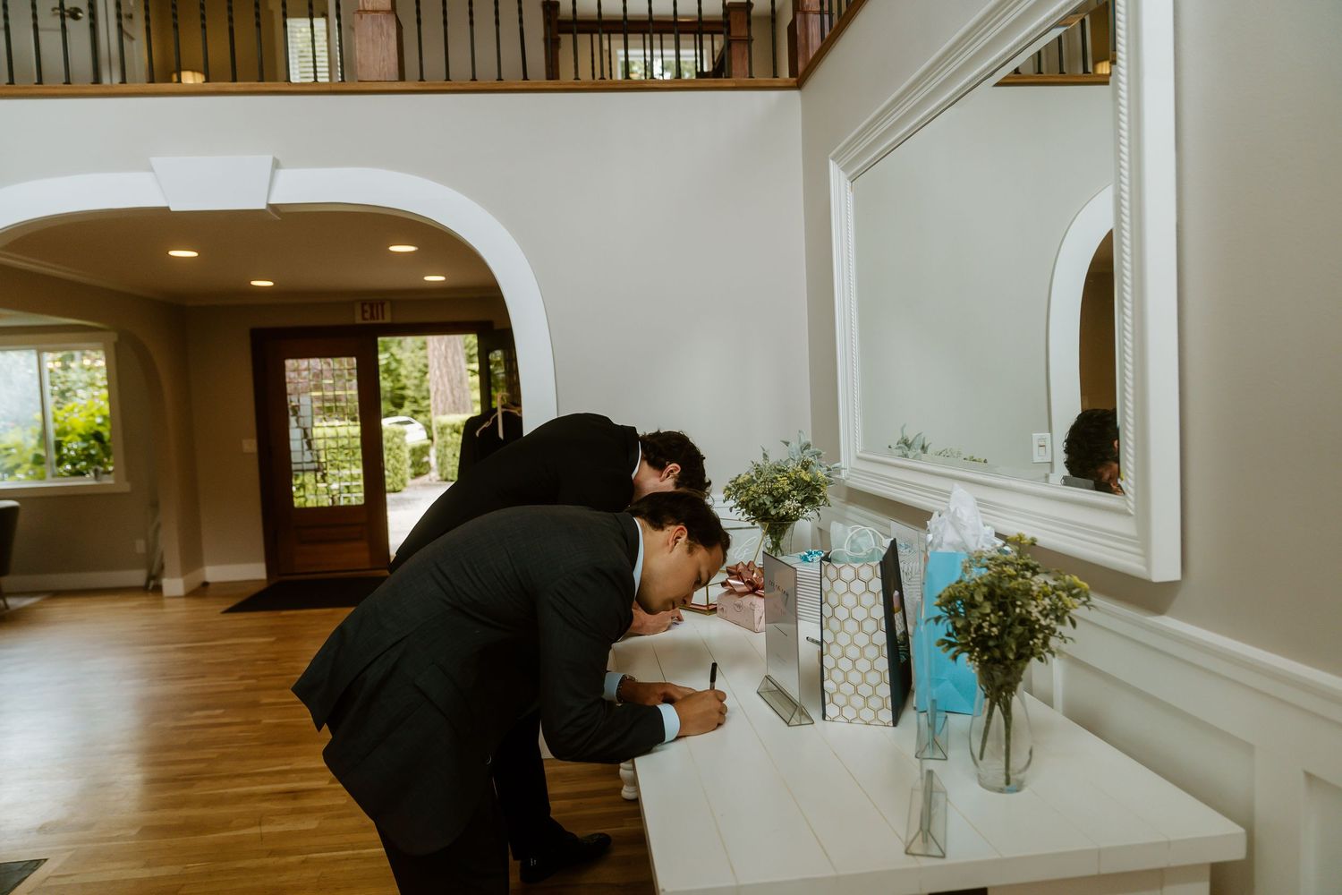 Two groomsmen in tuxedos signing the guest book at a wedding reception entrance table.