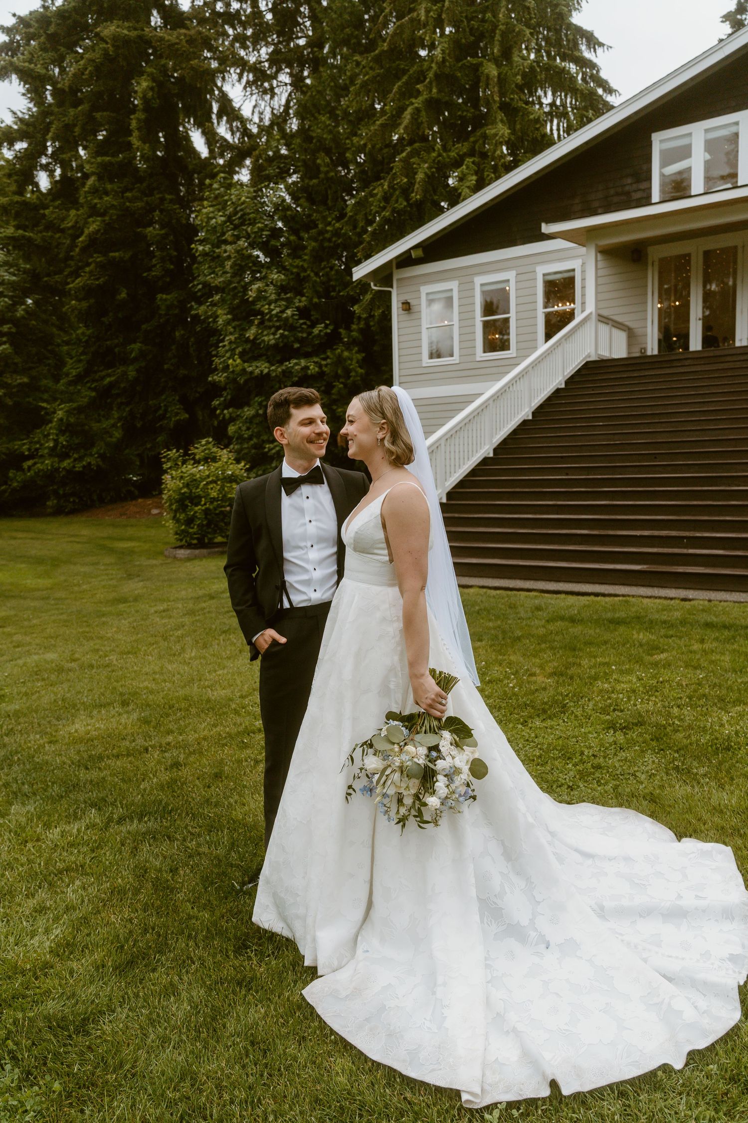 Wedding couple sharing a romantic moment on lush lawn beside a charming venue staircase surrounded by tall evergreen trees.