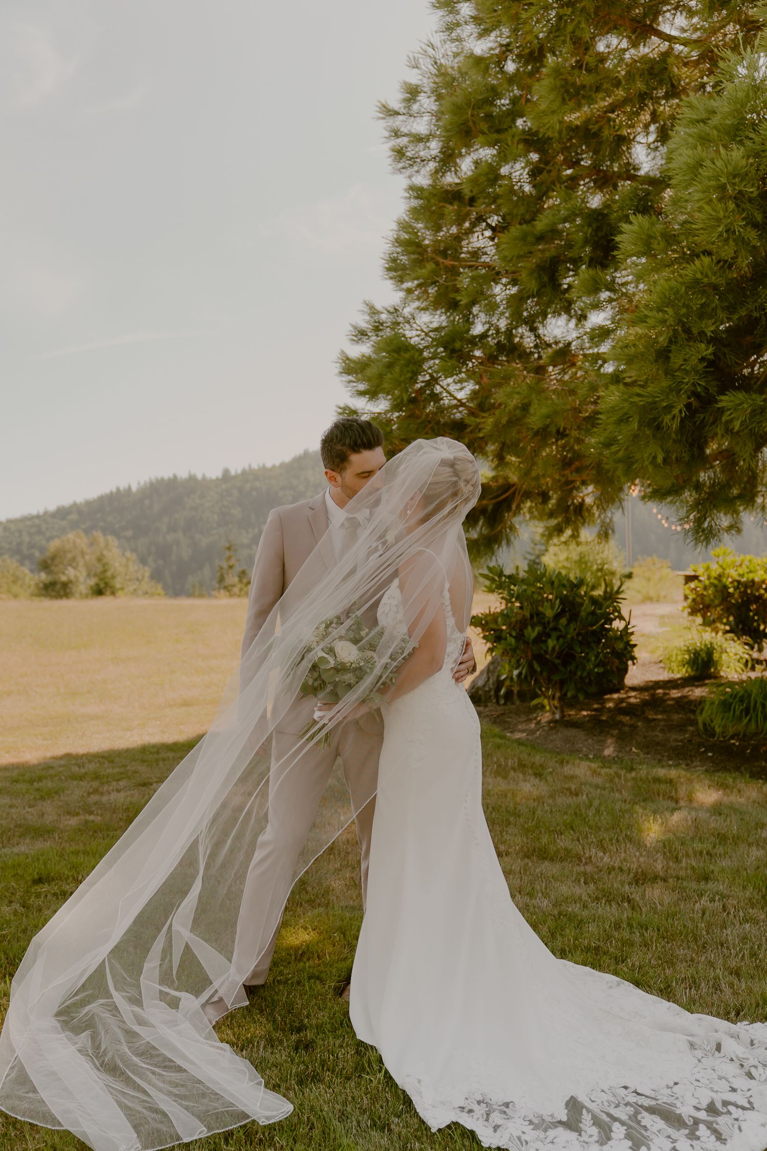 A flowing white wedding veil catches the breeze as a couple embraces in a scenic outdoor setting at sunset.