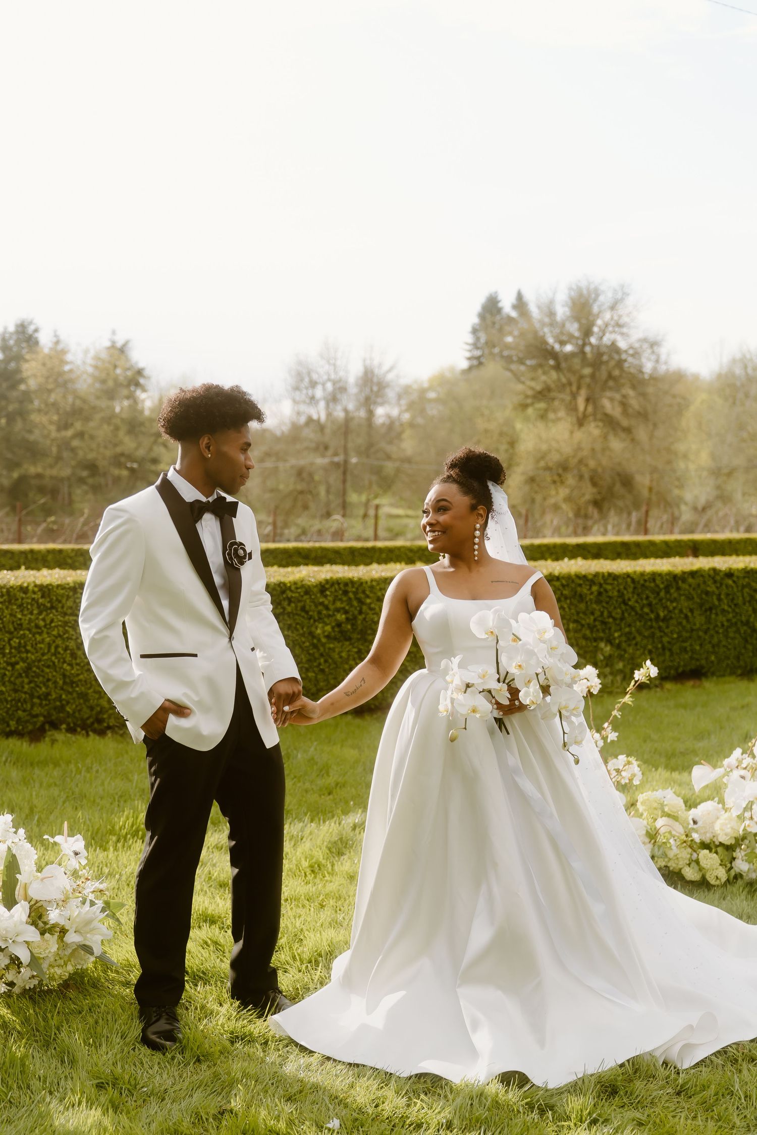 A Black bride and groom hold hands outdoors, she in a white ballgown with bouquet, he in a white tuxedo jacket.