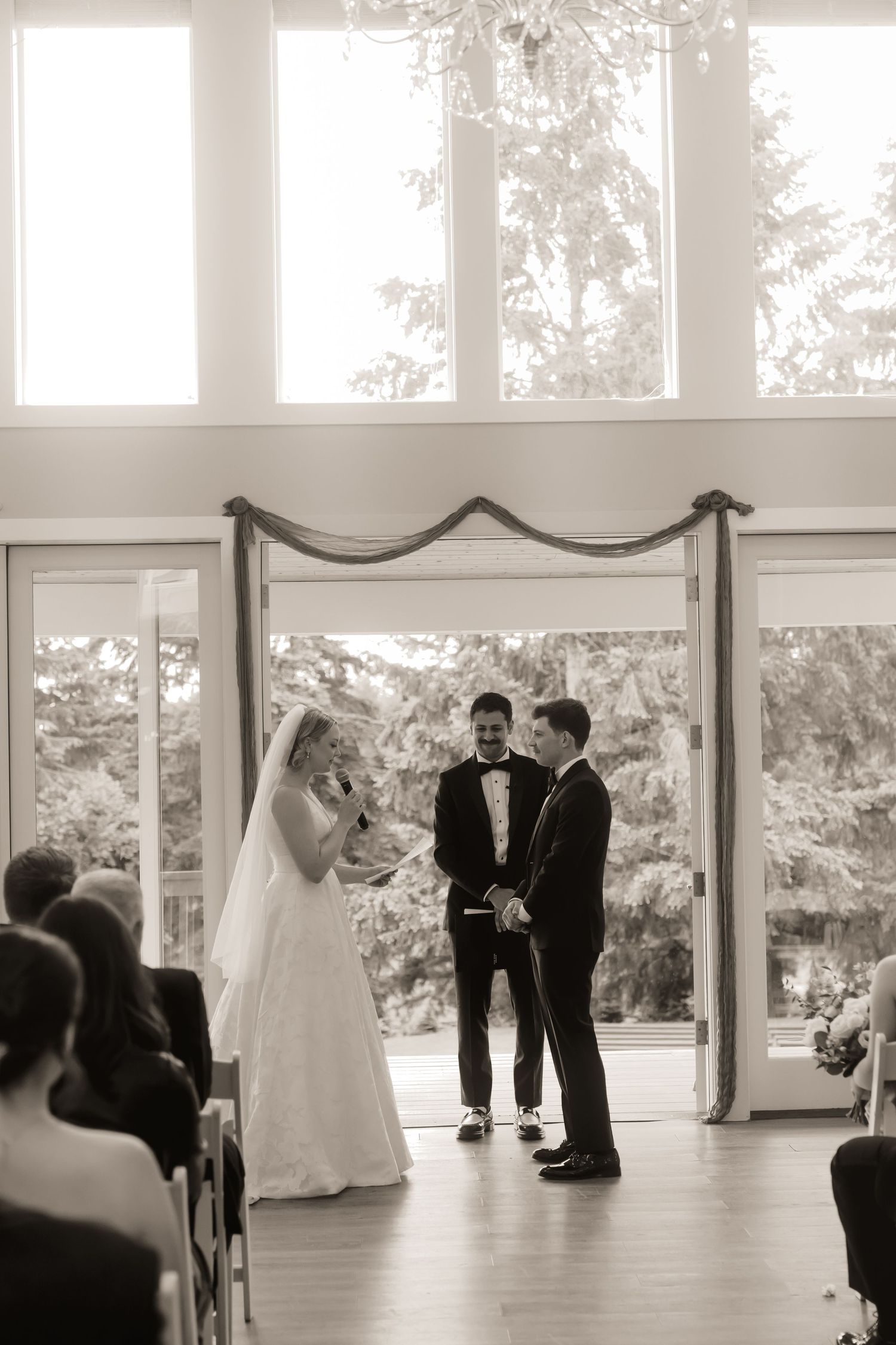 Black and white wedding ceremony photo of bride reading vows to groom before officiant, large windows with trees behind.