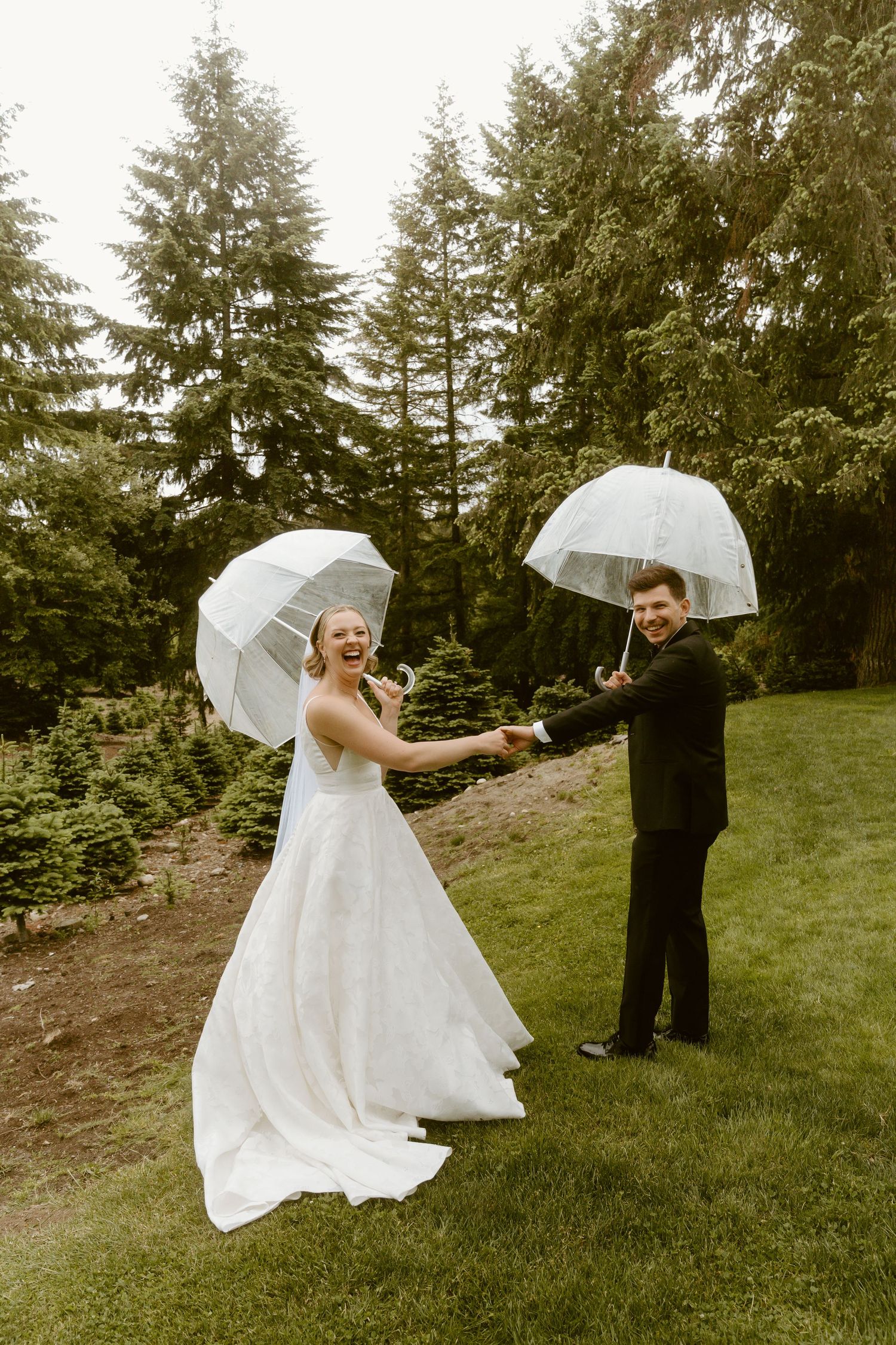Laughing bride and groom holding clear dome umbrellas on a rainy wedding day in a green outdoor setting.