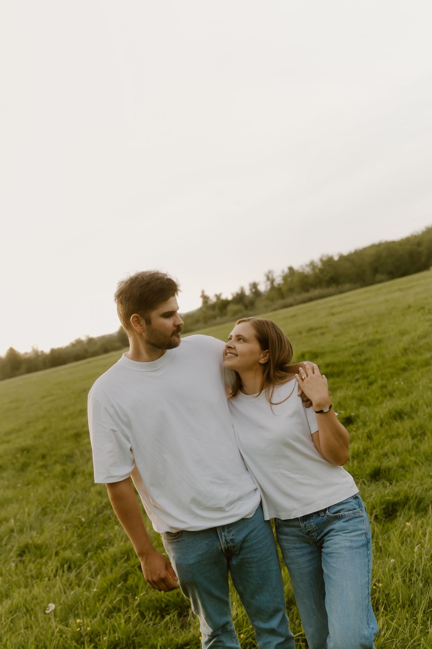 Couple in matching white t-shirts and jeans walking arm in arm through a green meadow at sunset.