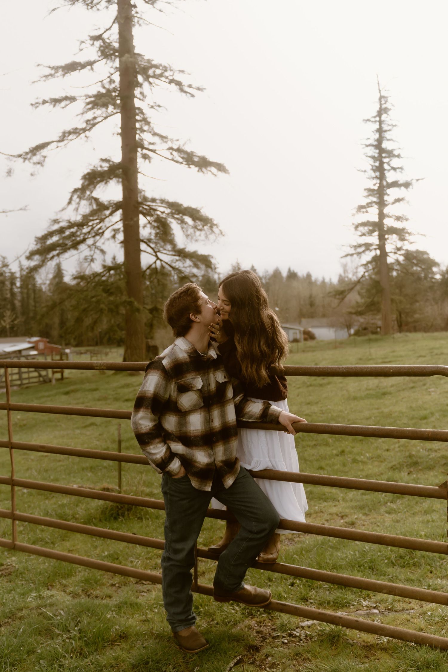 Couple sharing a romantic kiss leaning against a rustic farm fence surrounded by tall evergreen trees on a cloudy day.
