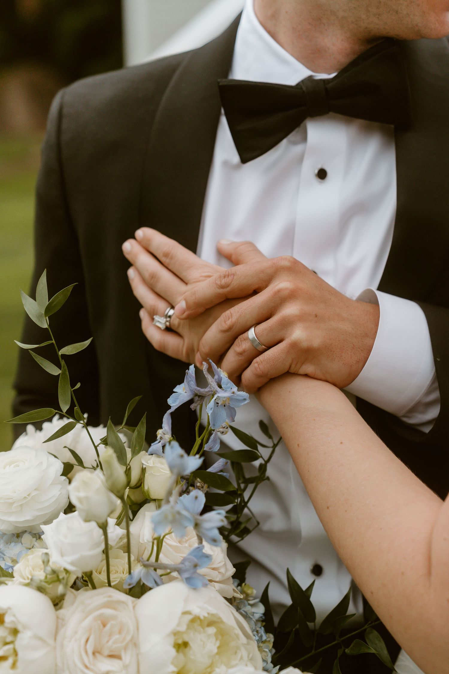 Bride and groom holding hands showing wedding rings with a white and blue floral bouquet.