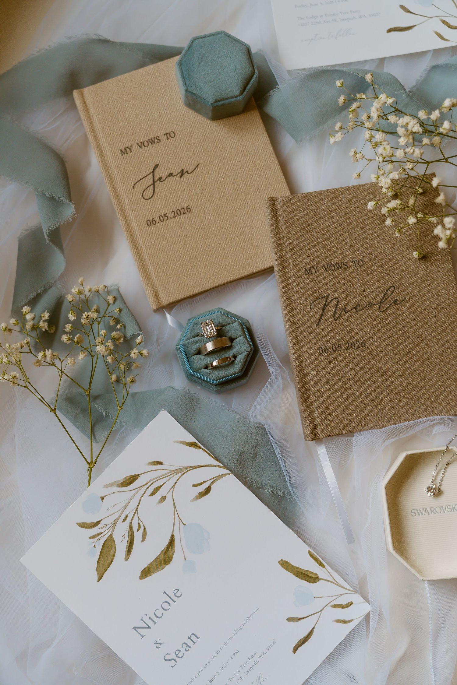 Elegant wedding flat lay with vow books, engagement rings in blue velvet box, floral invitation, and baby's breath on sheer fabric.