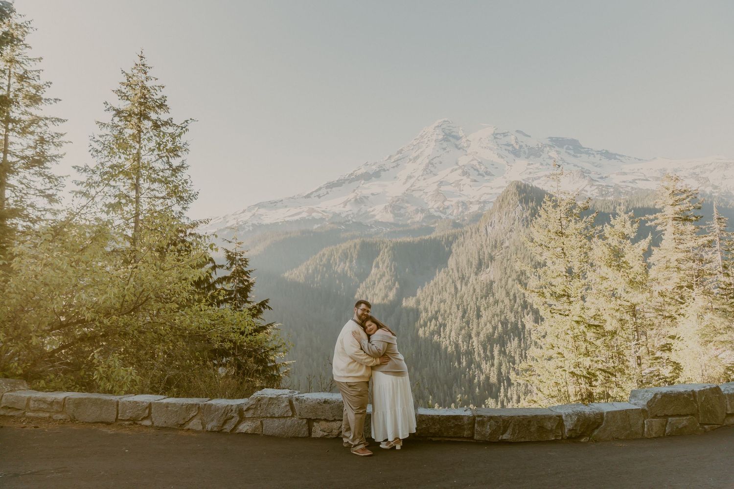 Couple embracing at a scenic mountain overlook with snow-capped peaks and pine trees in the background.