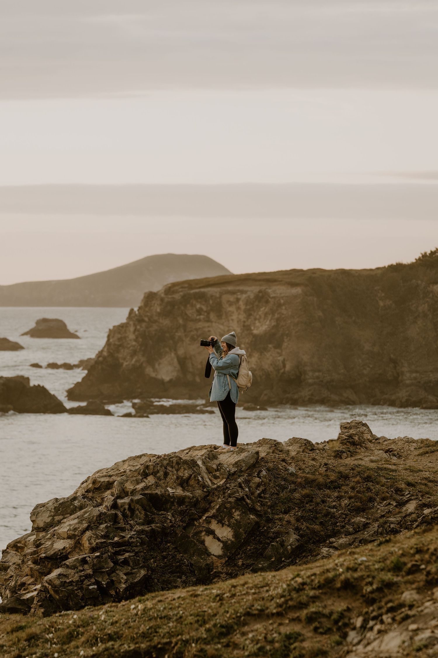 A photographer stands on rugged coastal cliffs at sunset, capturing the dramatic seascape with rocky outcrops and distant hills.