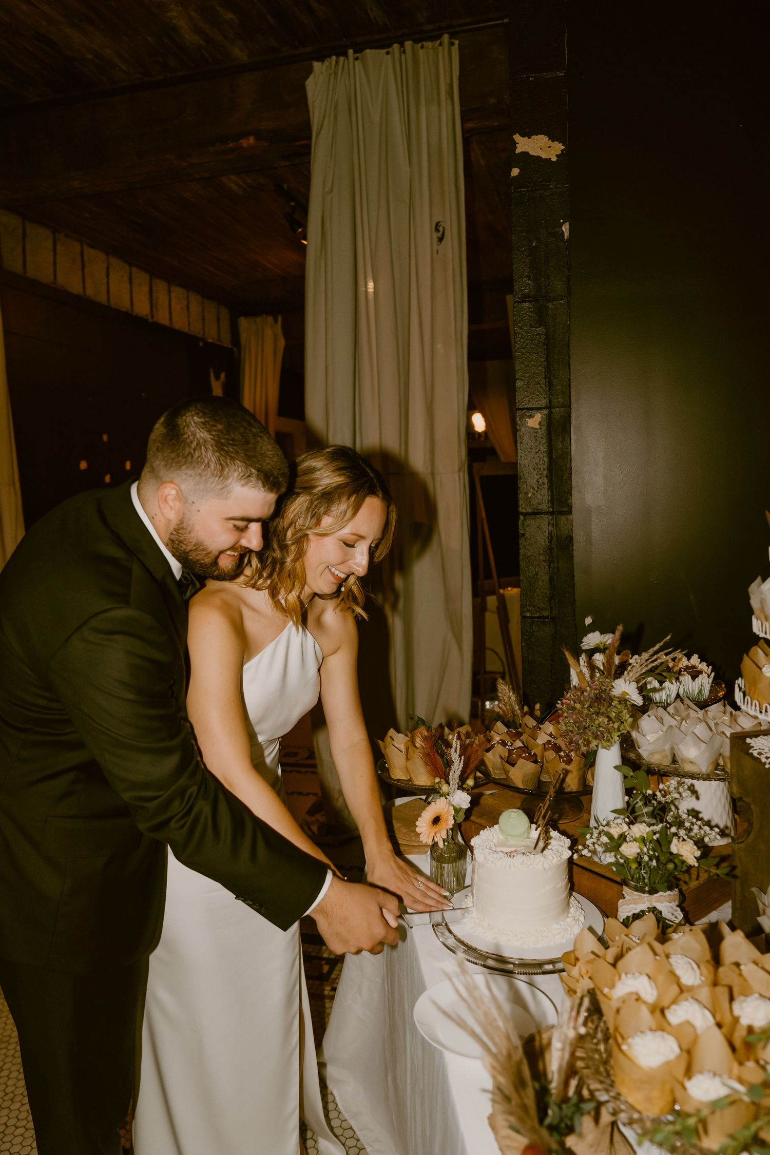 A couple cuts their elegant white wedding cake together in a dimly lit reception venue.
