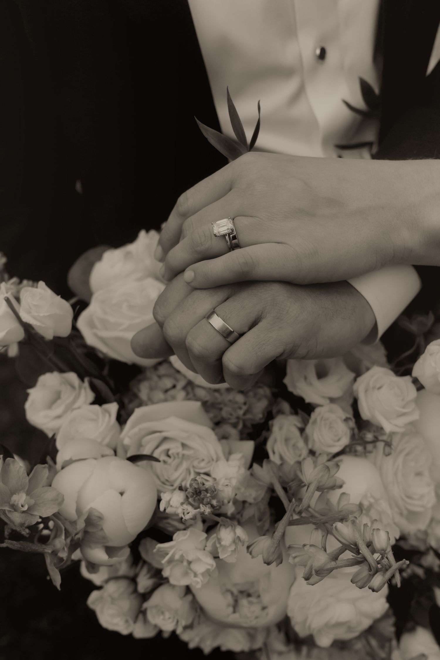 Black and white photo of newlyweds hands with wedding rings resting on a bouquet of roses.