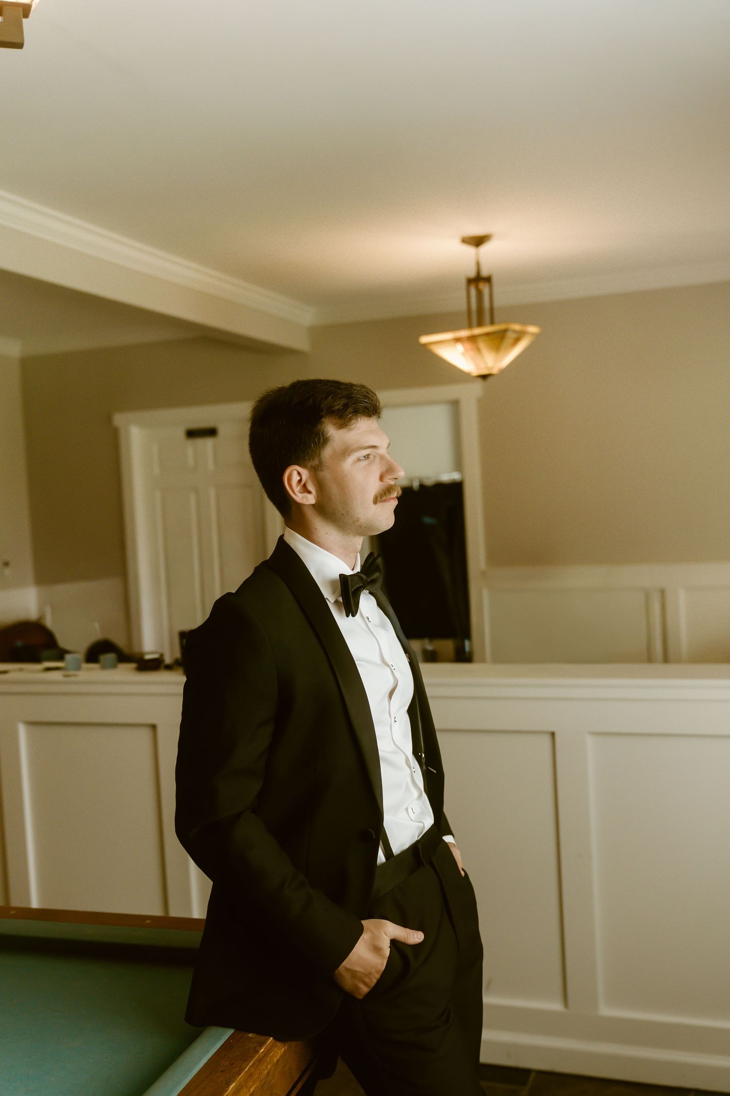Groom in black tuxedo and bow tie standing in a warmly lit room before wedding.