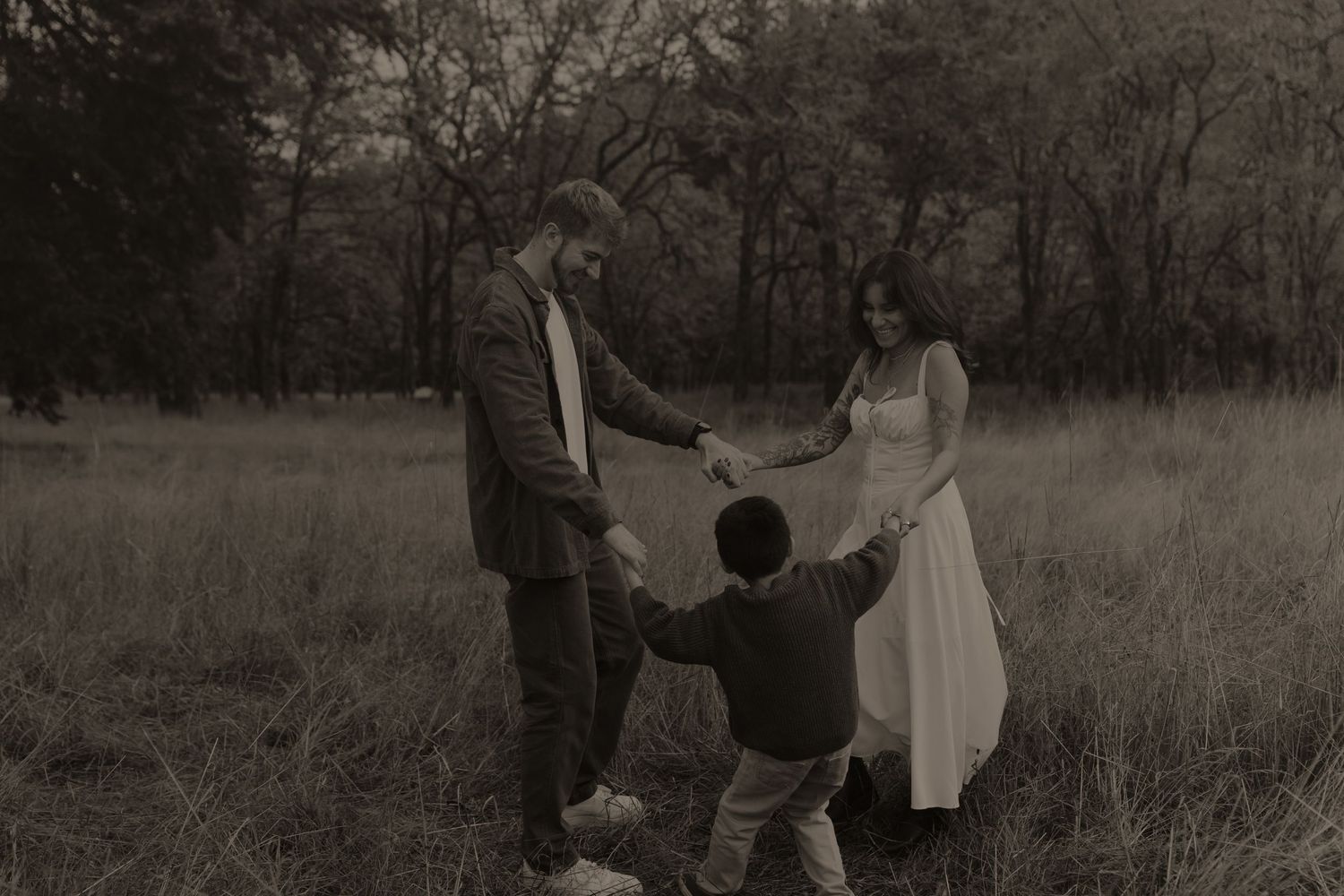 Family of three holding hands in a misty field, sepia-toned photo of parents and child playing outdoors.
